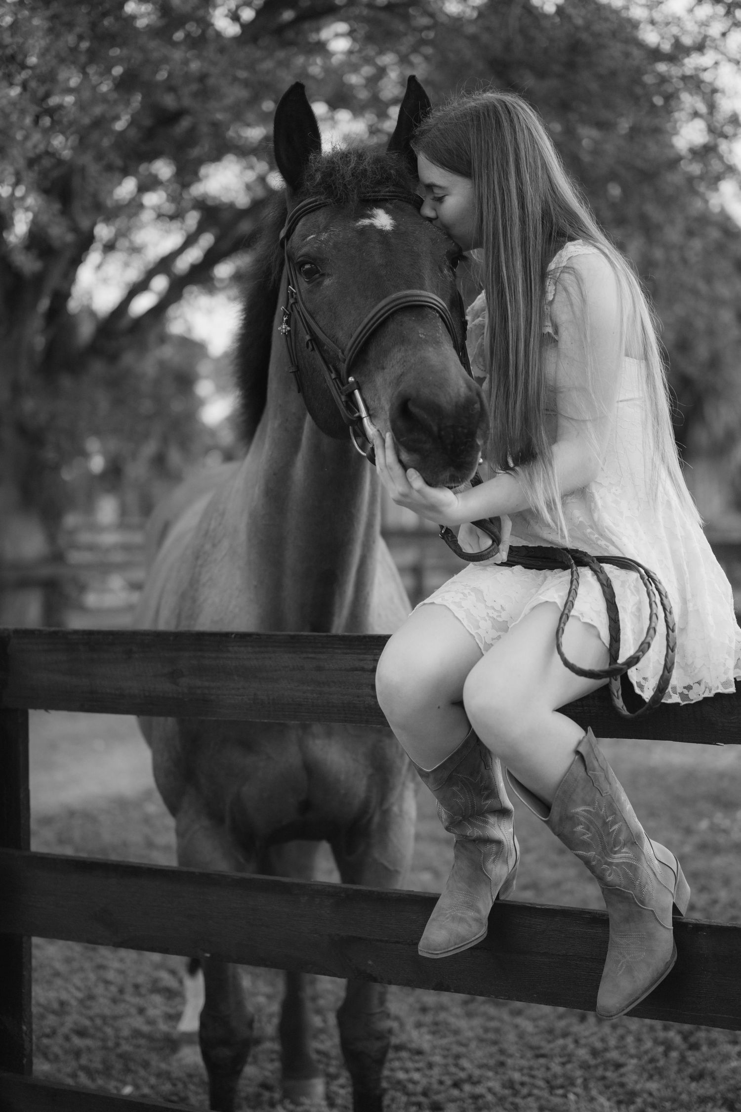Black and white portrait of young woman in cowboy boots sitting on fence kissing a bay horse.