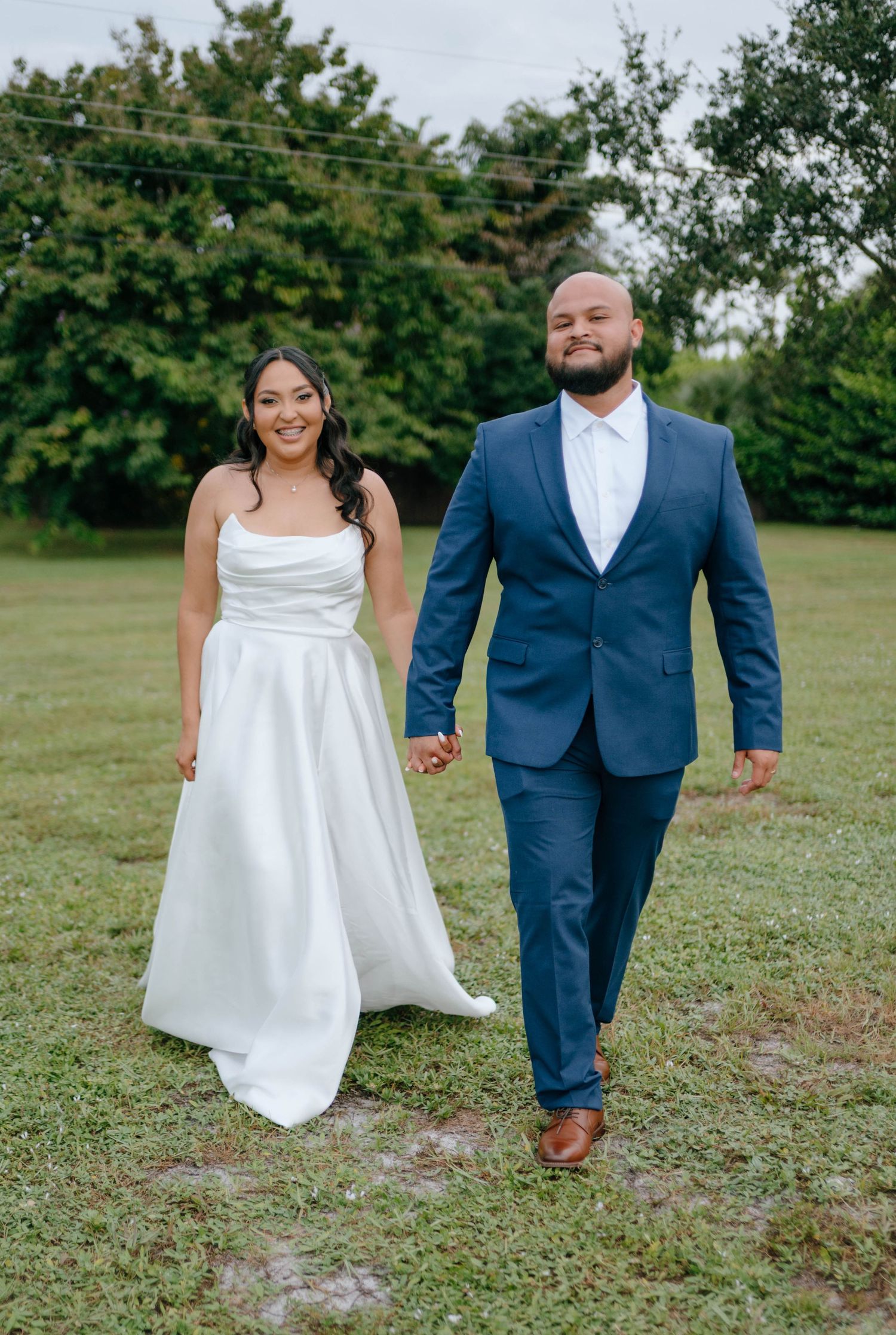 Bride in white satin gown and groom in navy blue suit hold hands walking outdoors on green grass.