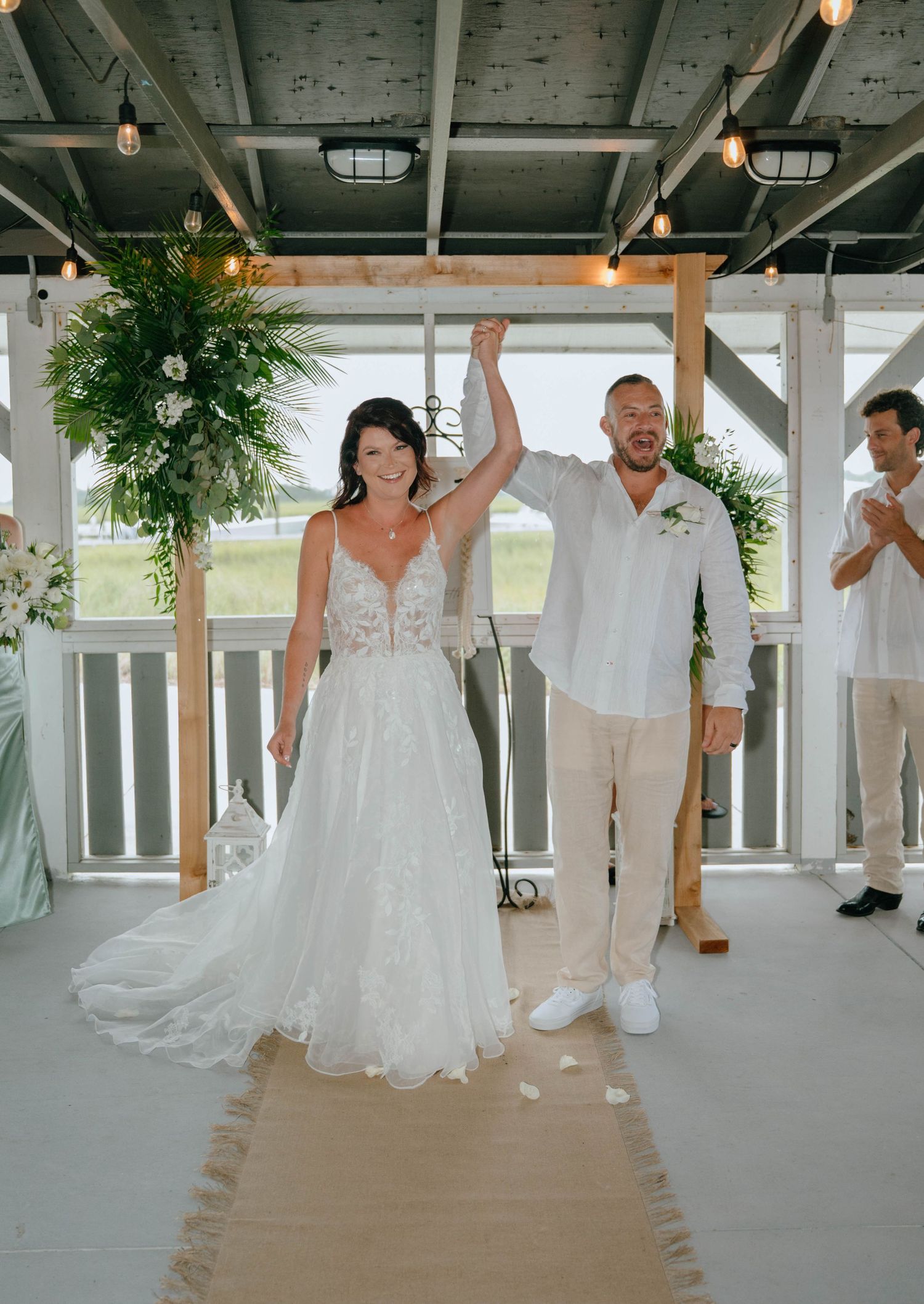 Newlywed couple raises joined hands in celebration after beach wedding ceremony on decorated porch.