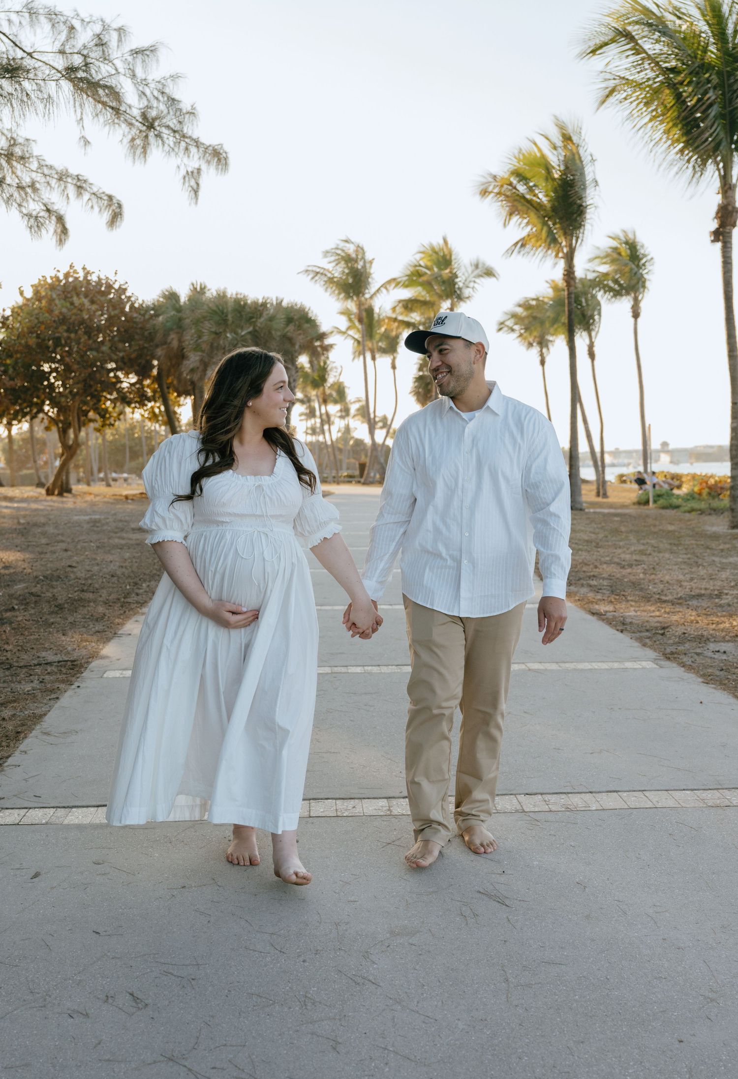 Happy expecting couple walks hand-in-hand on sunny palm tree path, both dressed in white.