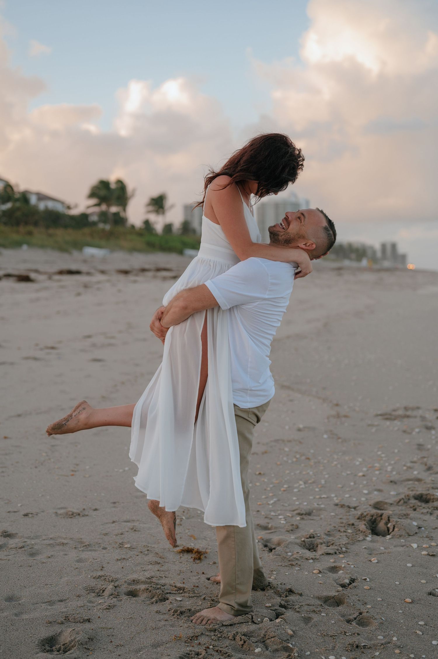 Man joyfully lifts woman in white dress on sandy beach at sunset during romantic photo session.