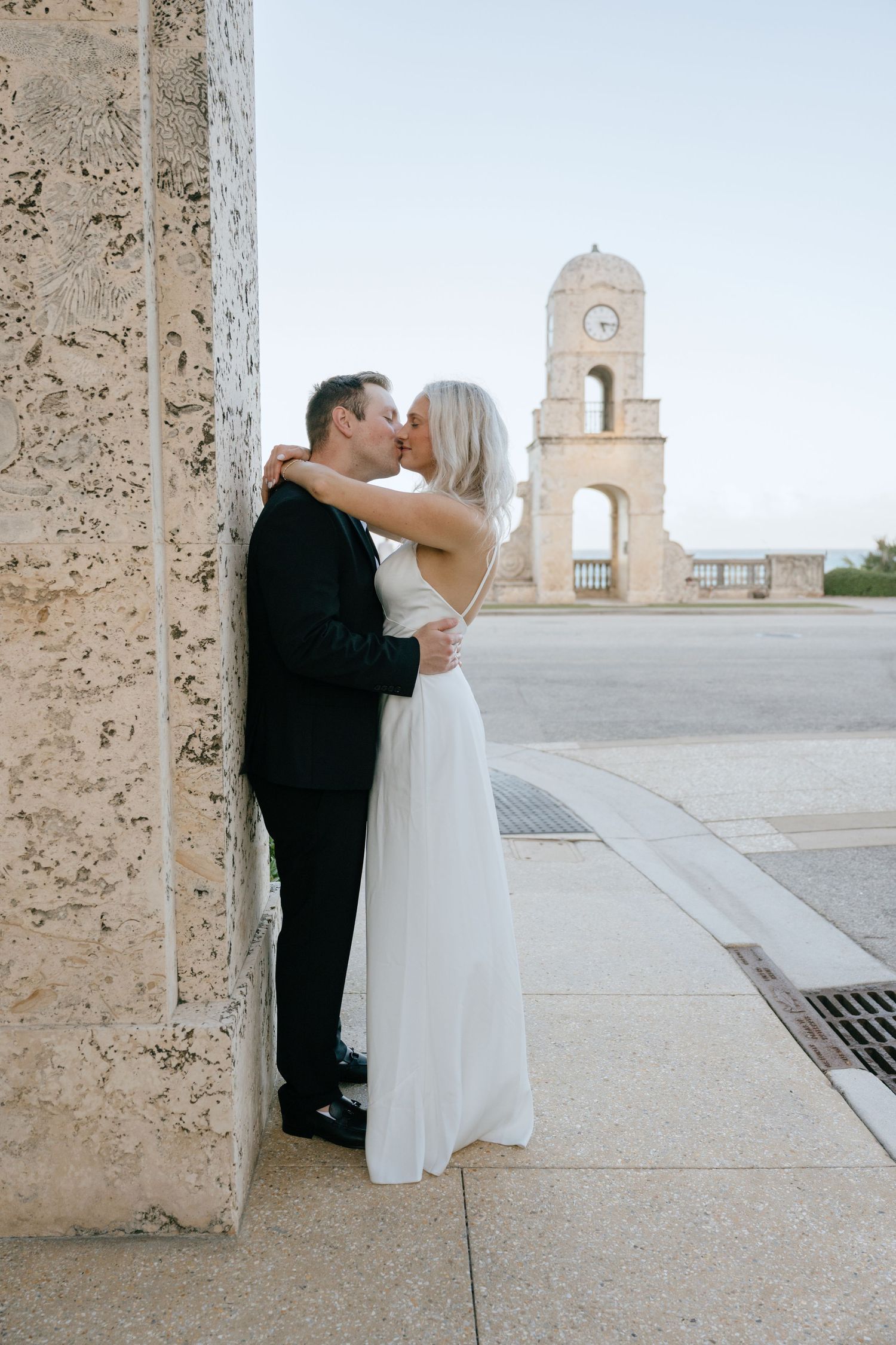 Newlyweds share a romantic kiss against a stone wall near a classic clock tower at dusk.