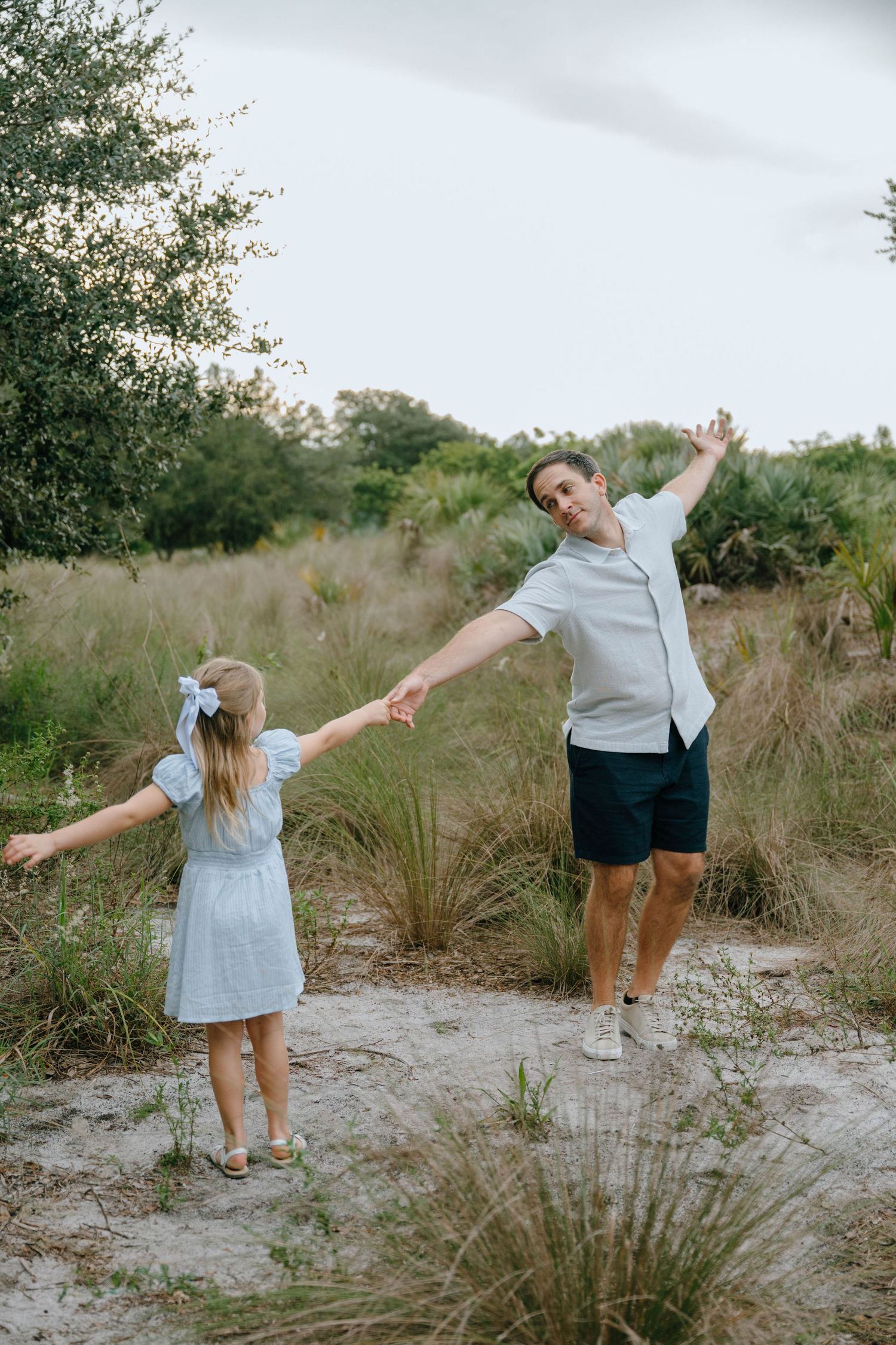A father and young daughter hold hands and dance playfully on a sandy path surrounded by greenery.