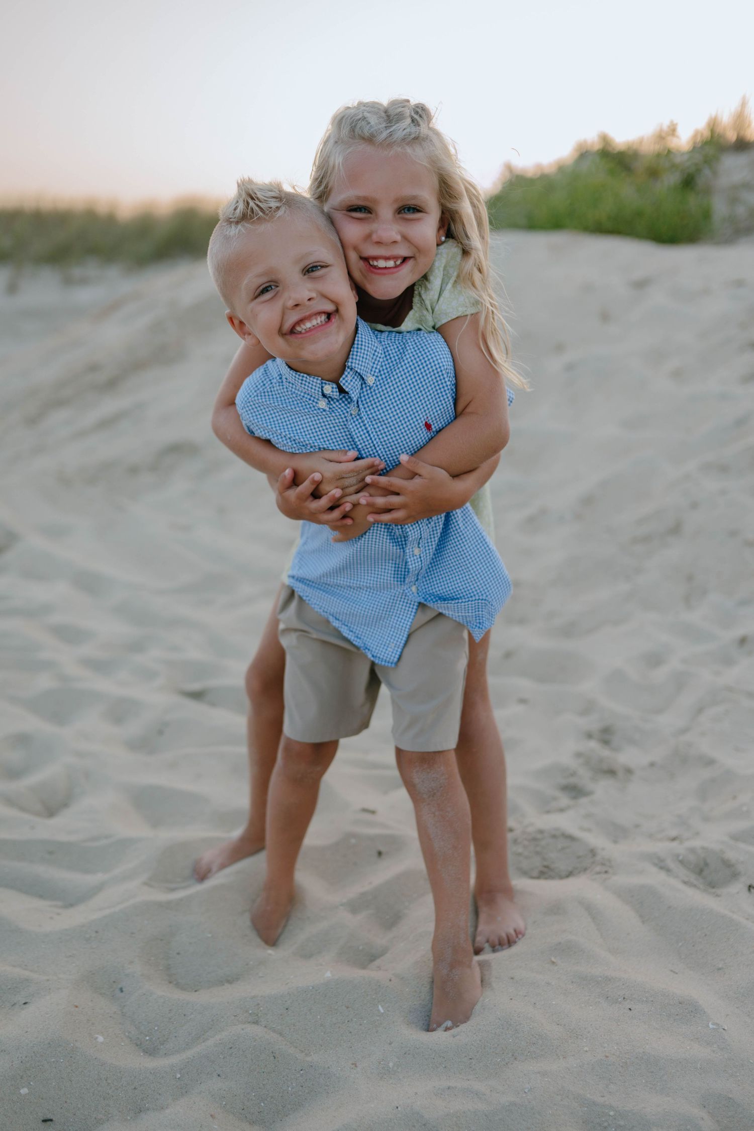 Two young siblings laugh and hug on sandy beach dunes at sunset, girl embracing boy from behind.
