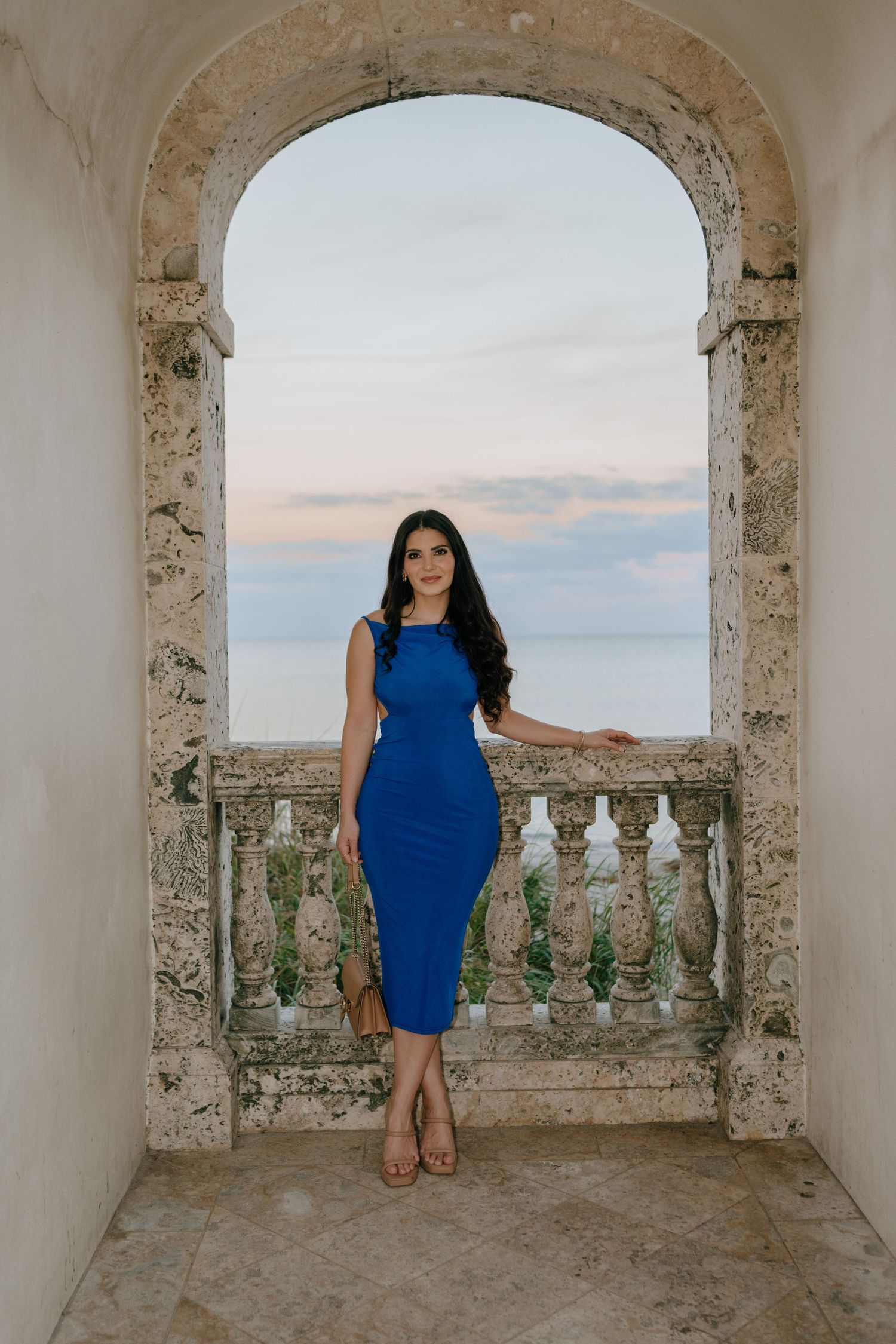 Woman in blue midi dress posing under stone arch with ocean view at sunset.