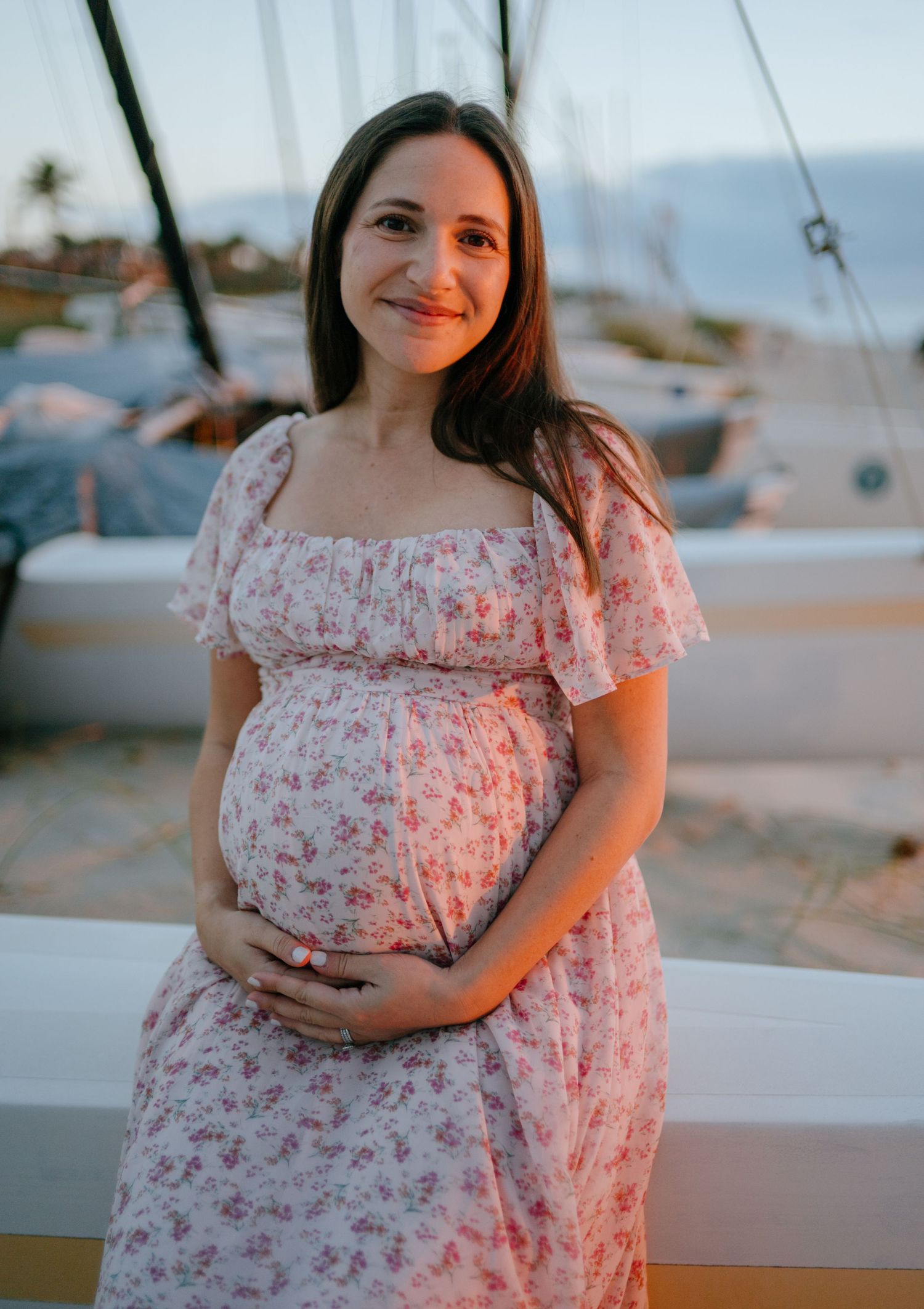 Smiling pregnant woman in floral dress cradling baby bump at sunset near sailboats.