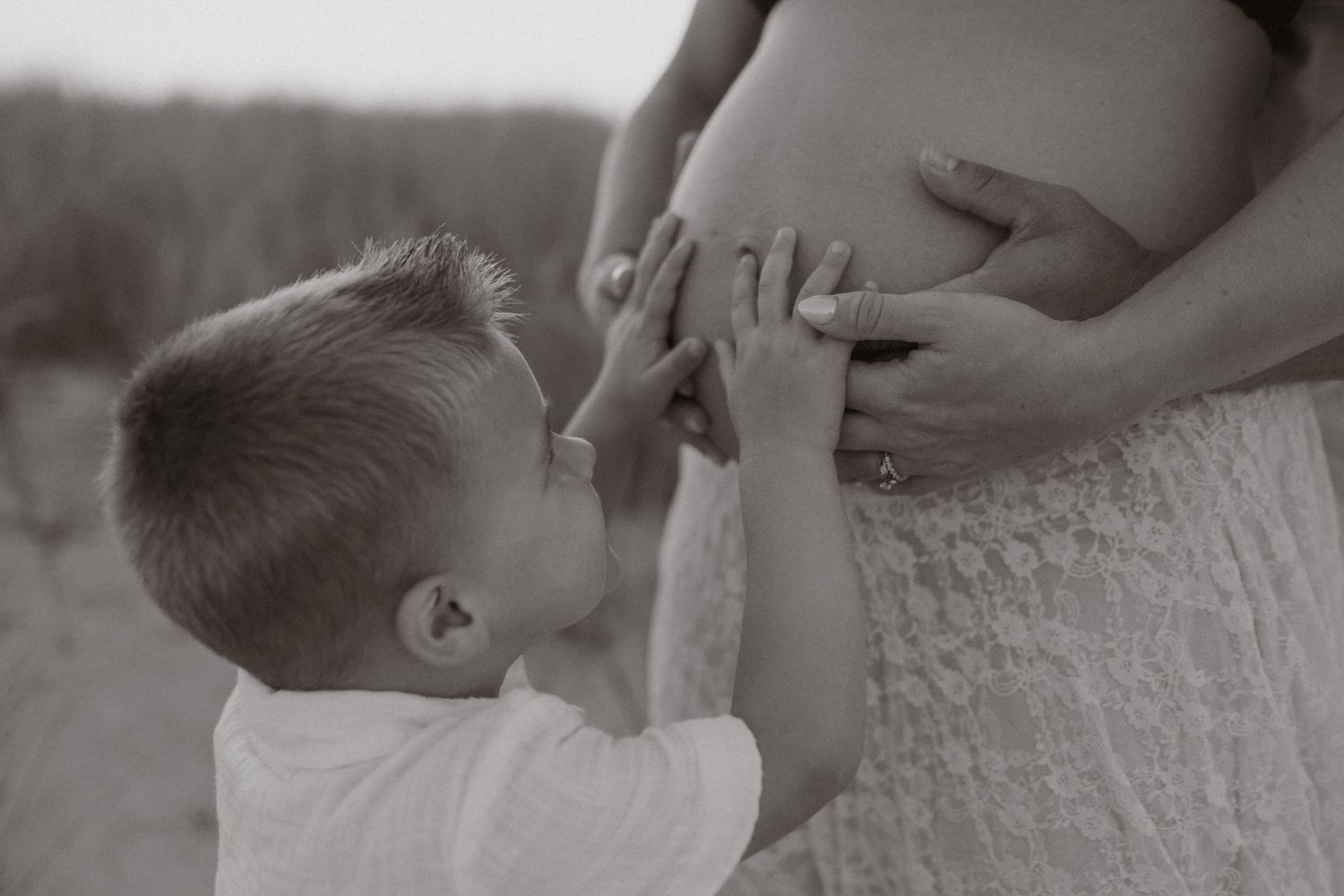 Black and white photo of a young boy kissing a pregnant woman's belly during a maternity photoshoot.