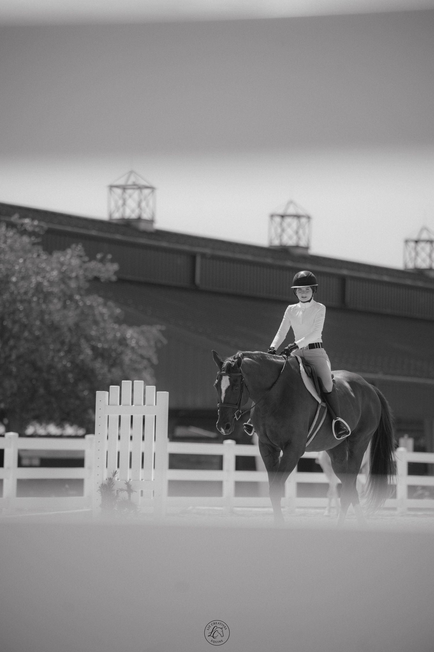 Black and white photo of equestrian rider on dark horse in sandy arena near white fence and building.