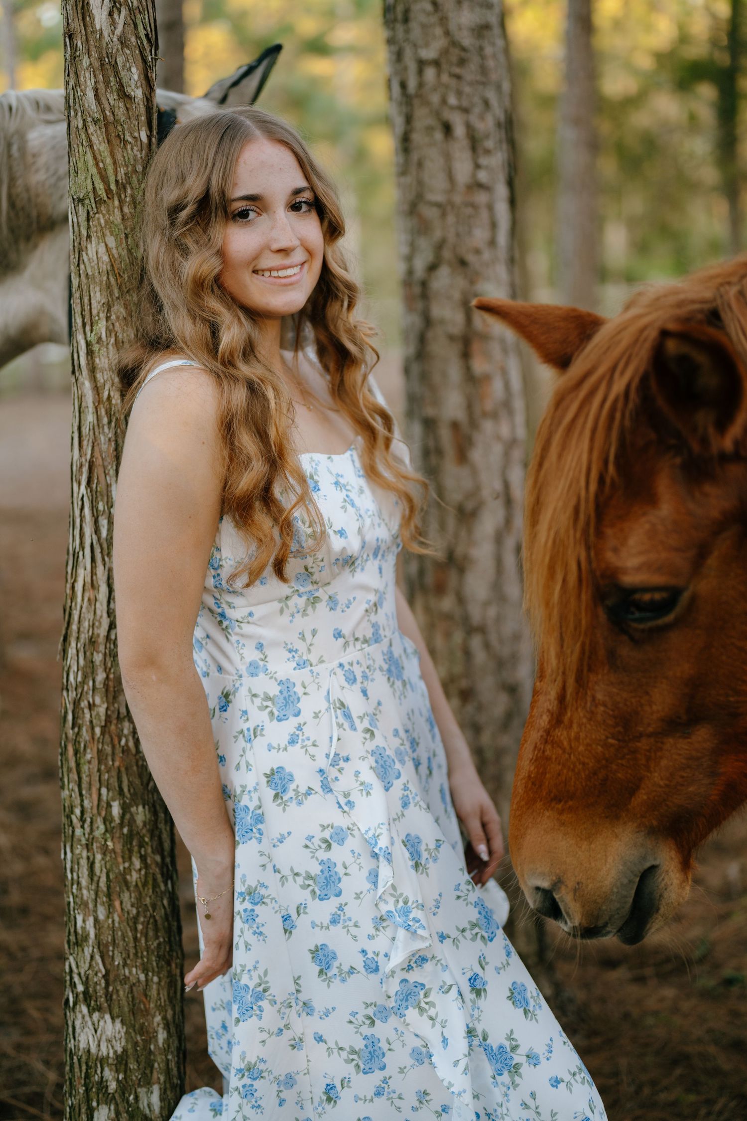 A young woman in a blue floral sundress leans against a tree while a chestnut horse grazes nearby.