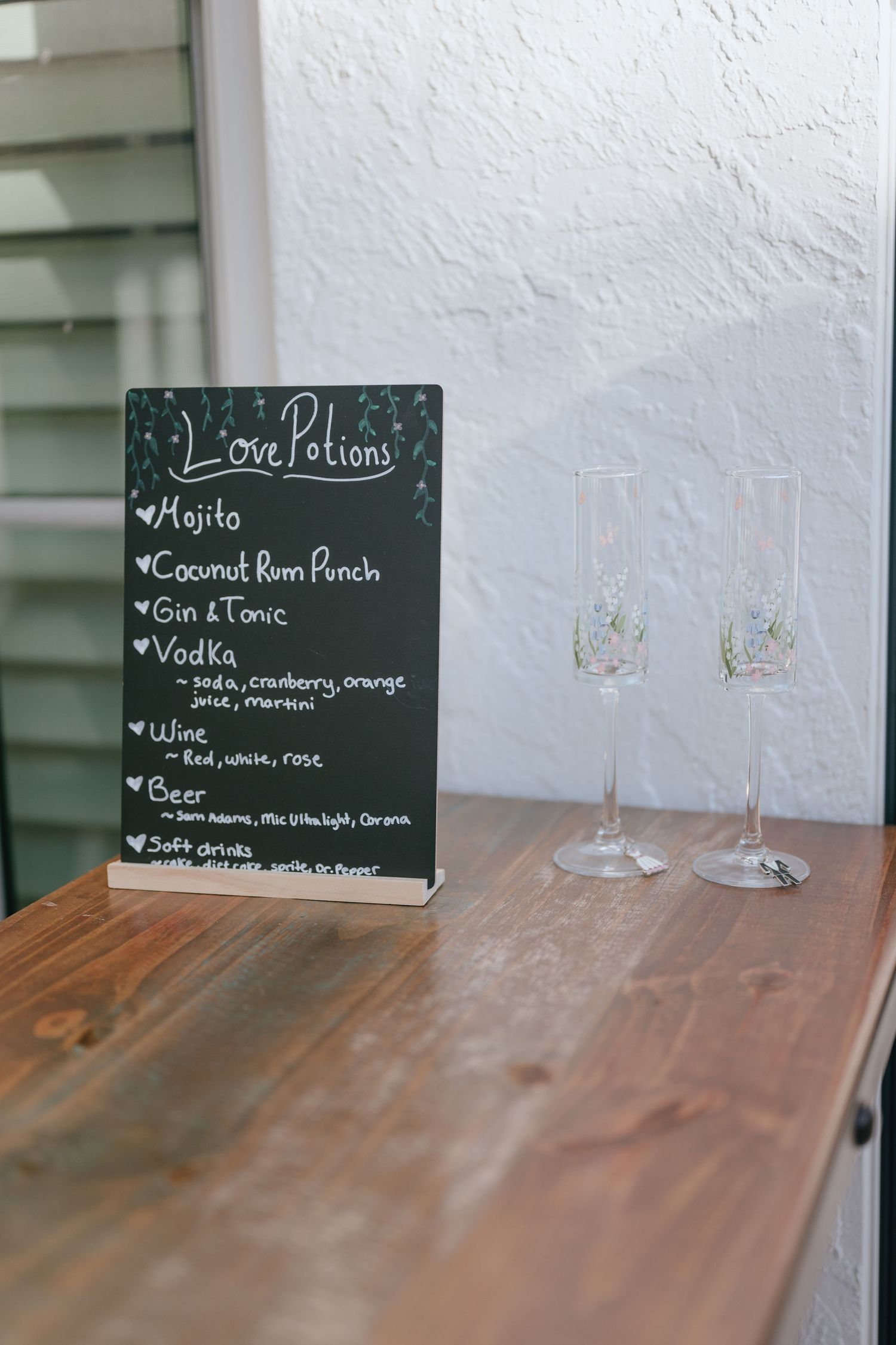 Wedding bar menu chalkboard sign listing love potions cocktails beside two champagne flute glasses.