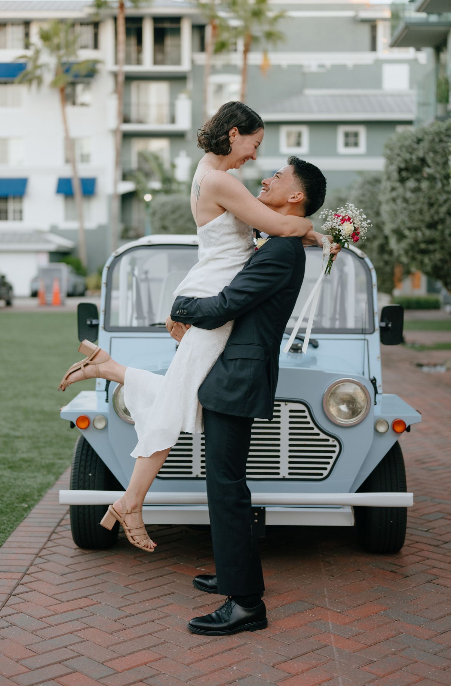 Groom lifting smiling bride in front of a vintage light blue Moke car on a brick courtyard.