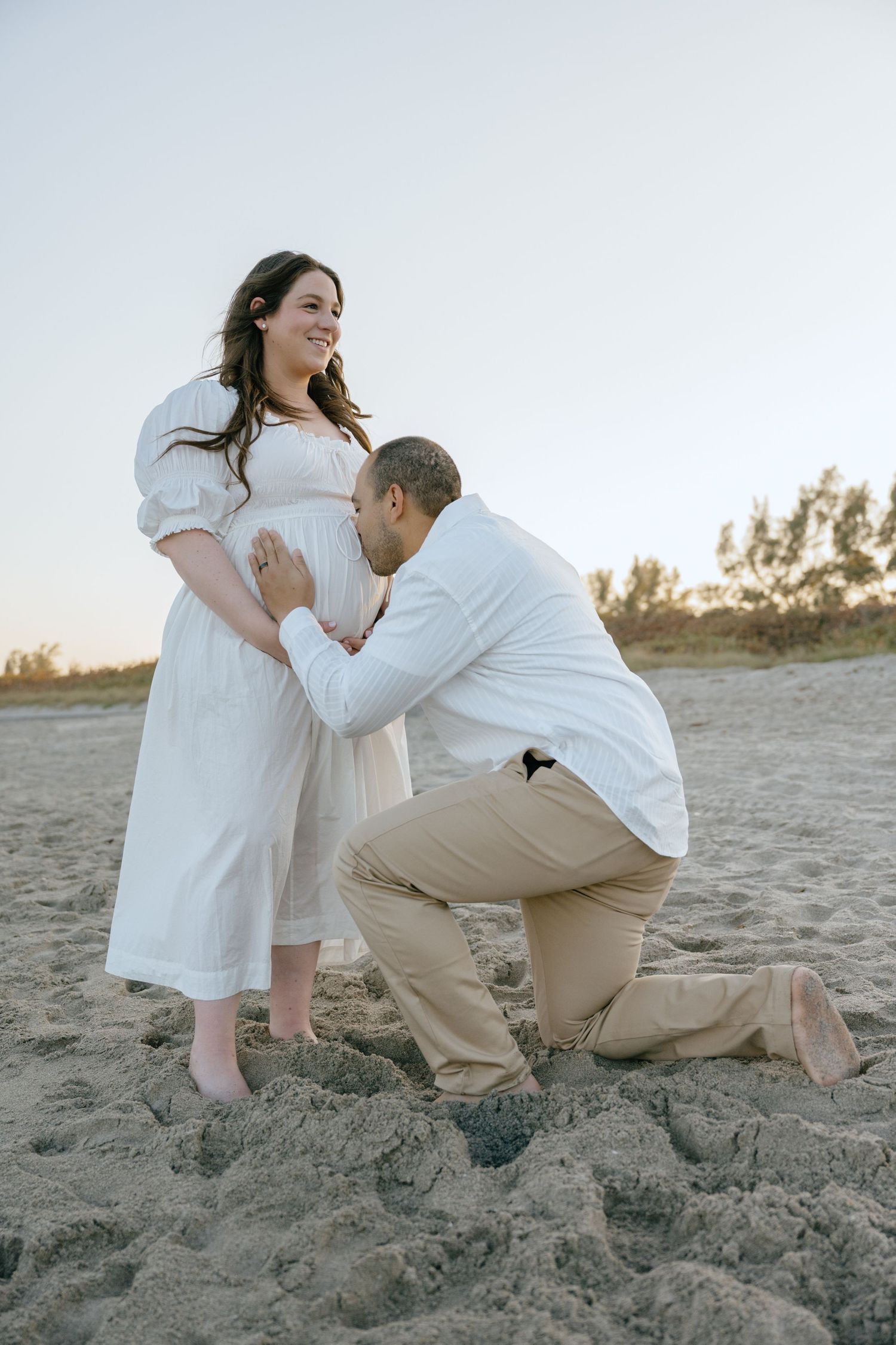 Man kneels in sand to kiss pregnant partner's baby bump on a beach at sunset.