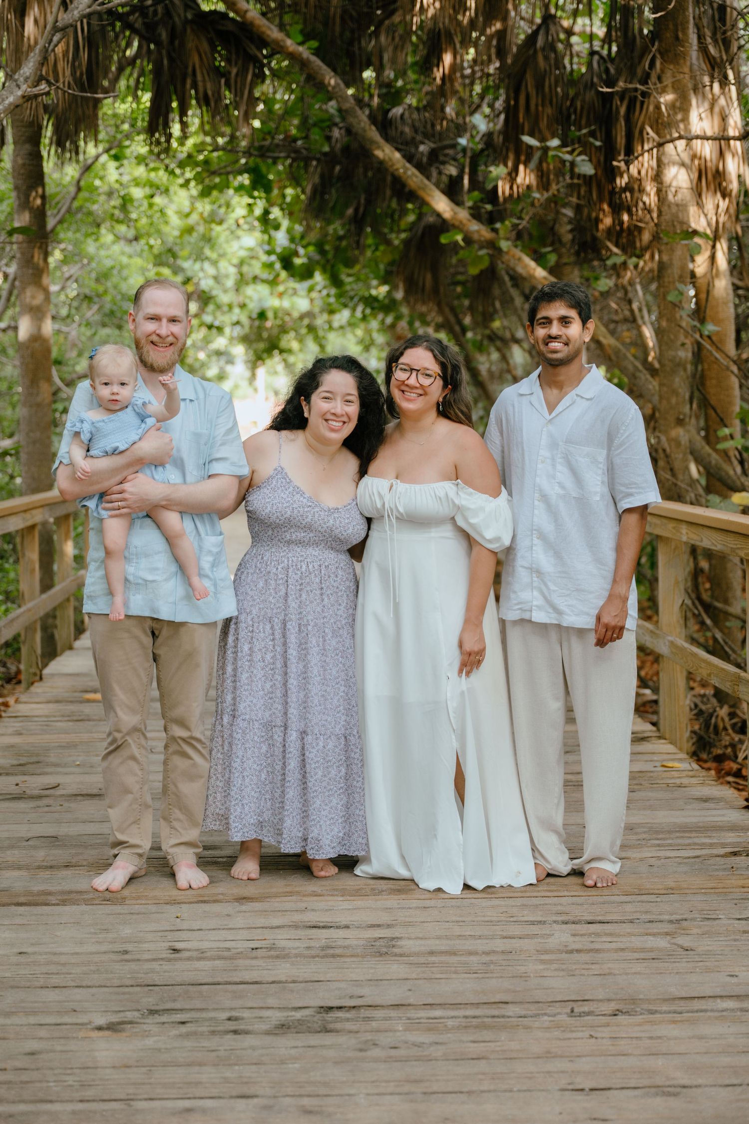 Five adults and a baby pose together smiling on a wooden bridge surrounded by lush green trees.