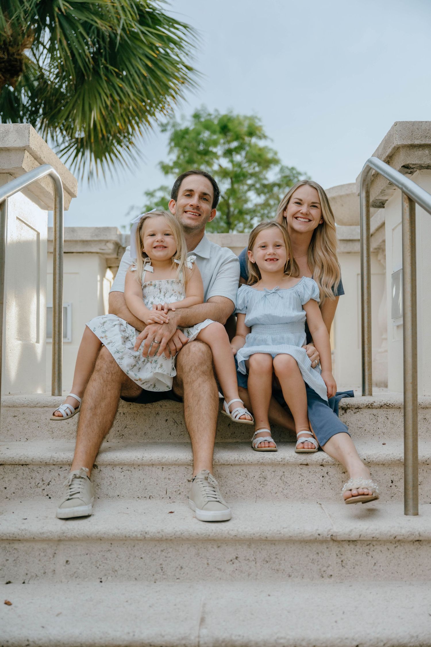 A happy family of four sits together on outdoor stone steps beneath a palm tree.