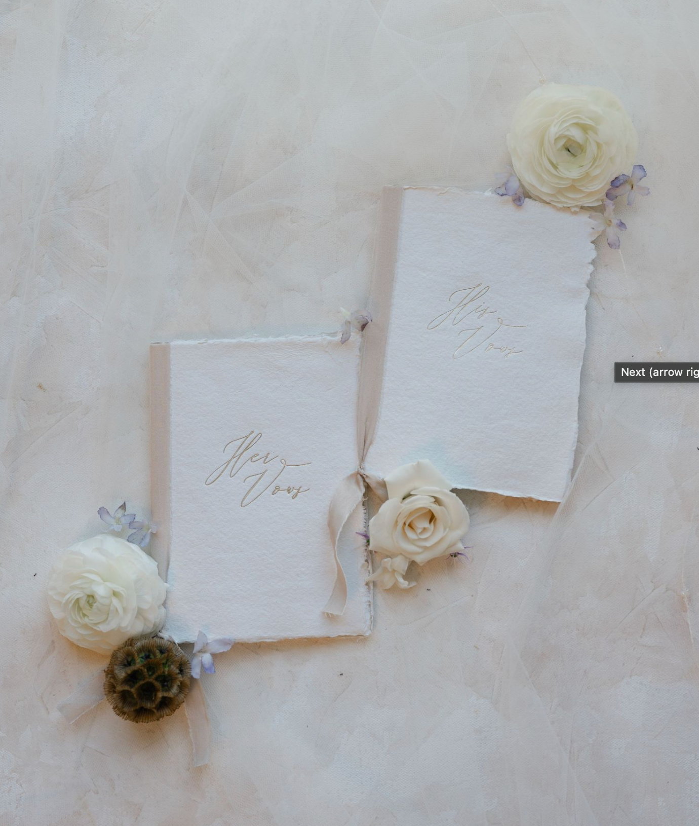 Elegant wedding vow books with gold calligraphy, adorned with white ranunculus and soft blue flowers on a marble surface.