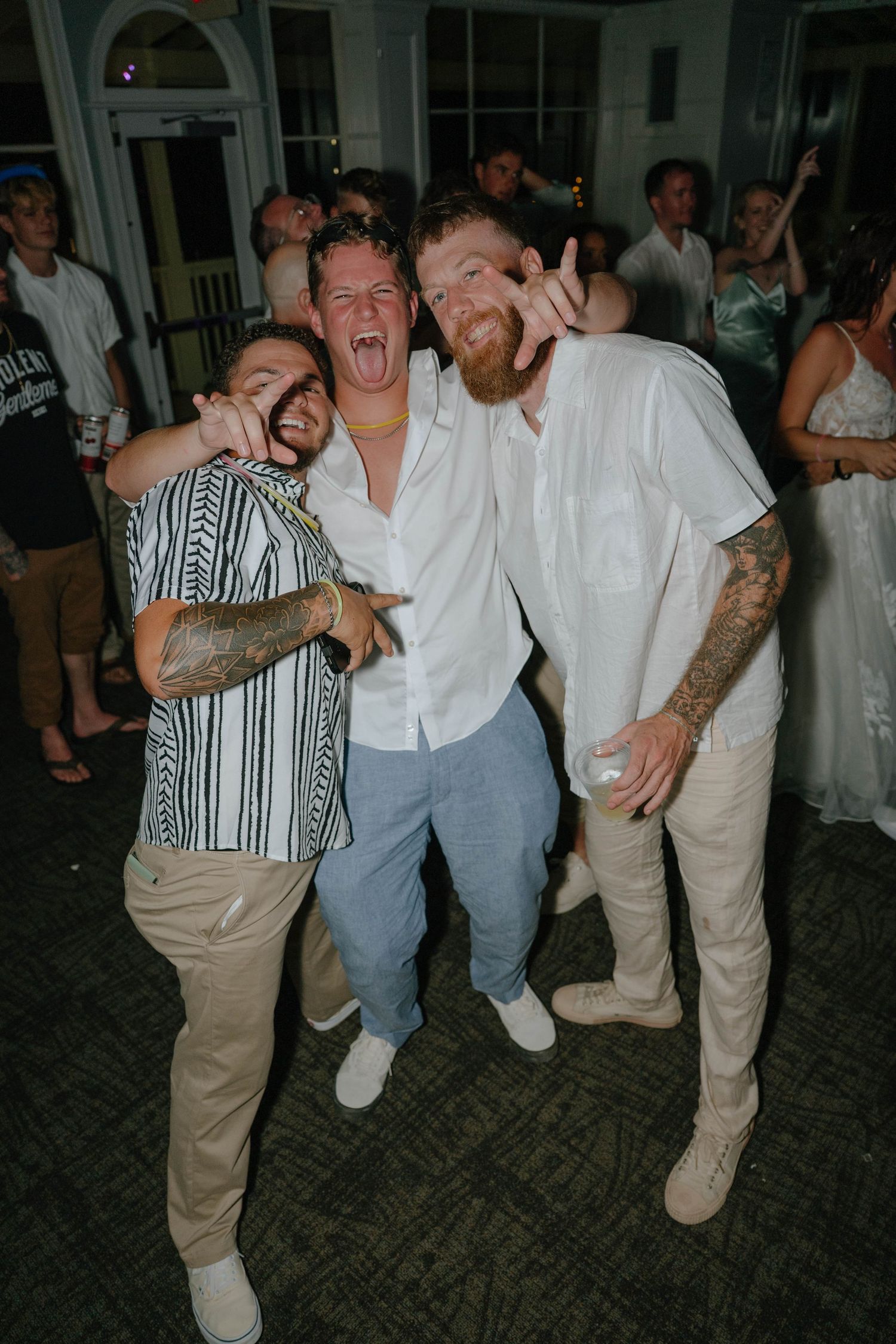 Three men in casual white and patterned shirts pose and smile together at an outdoor wedding reception.