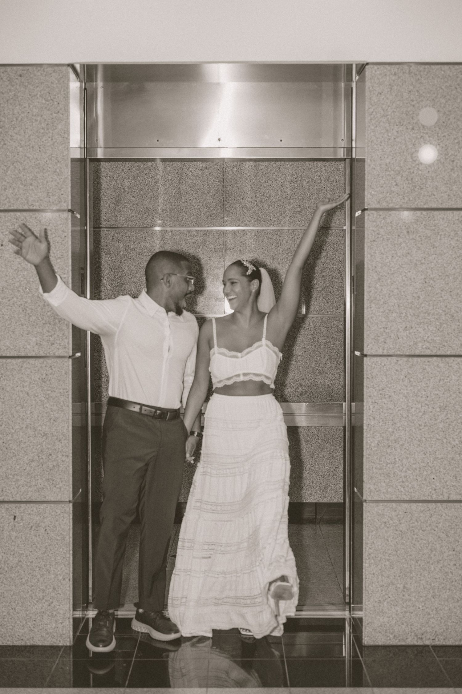 Black and white photo of bride and groom dancing joyfully inside a glass elevator.