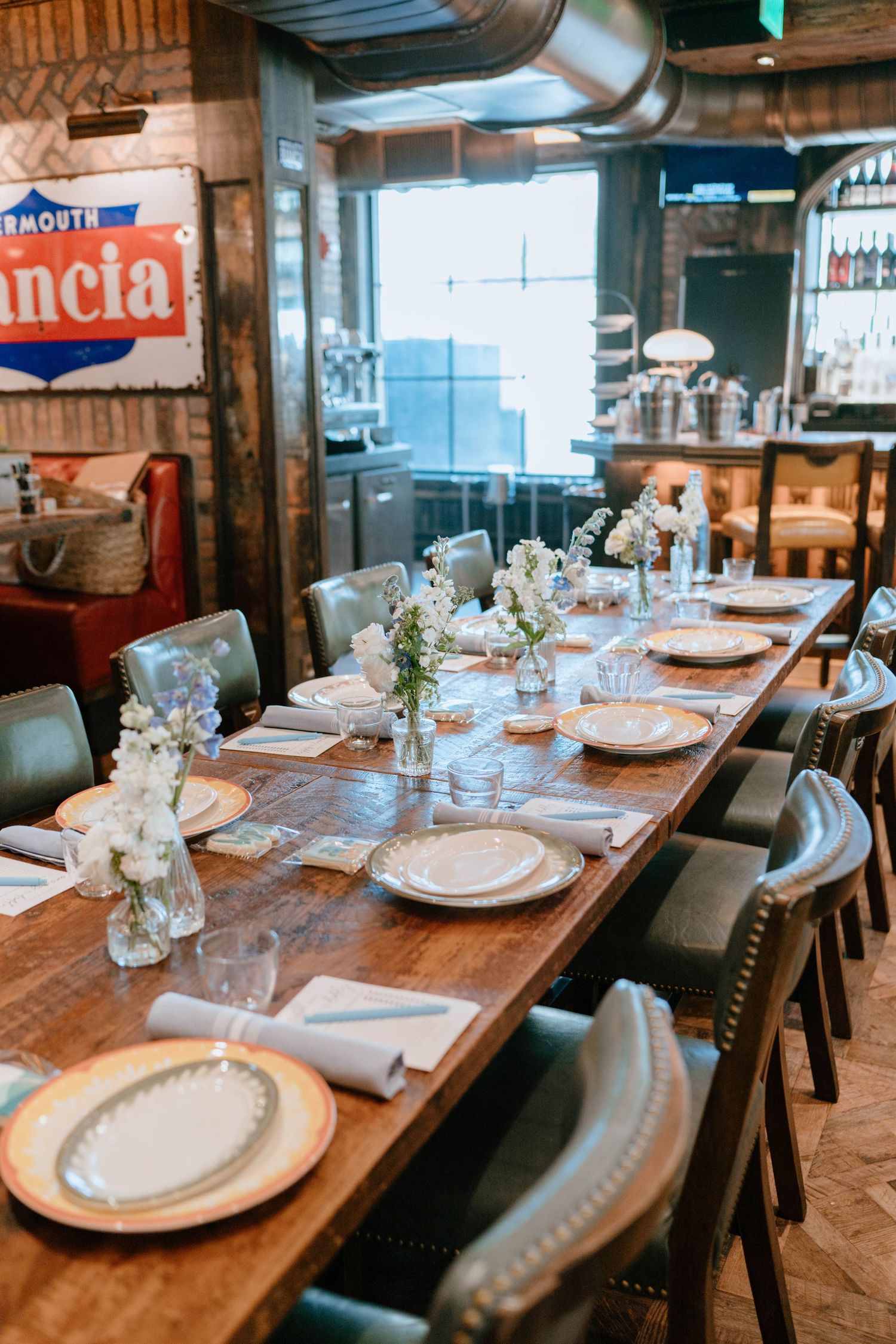 Rustic restaurant private dining table set with white plates, floral centerpieces, and leather chairs for a baby shower.
