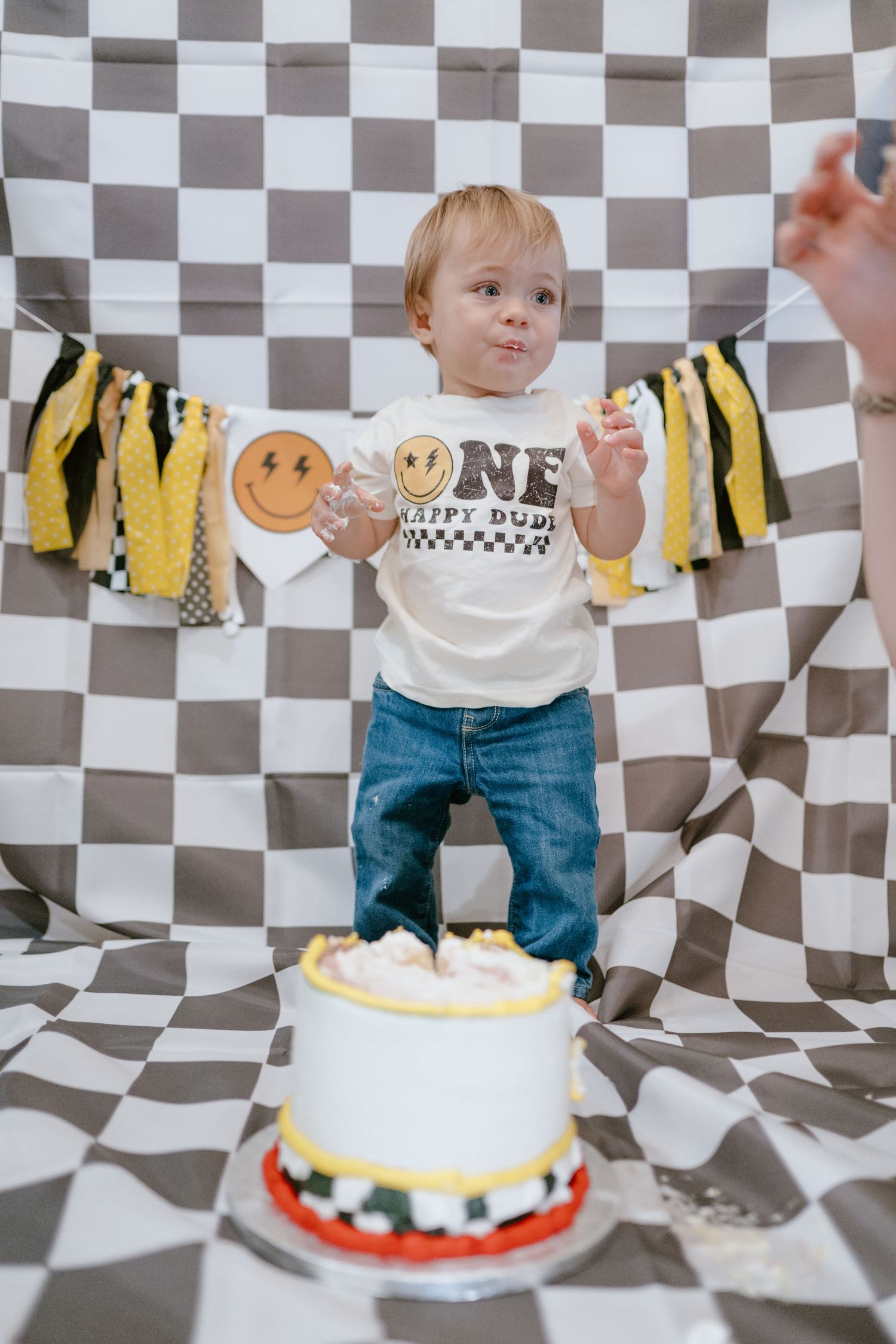 Standing toddler in ONE smiley face shirt poses with first birthday smash cake on checkered backdrop.