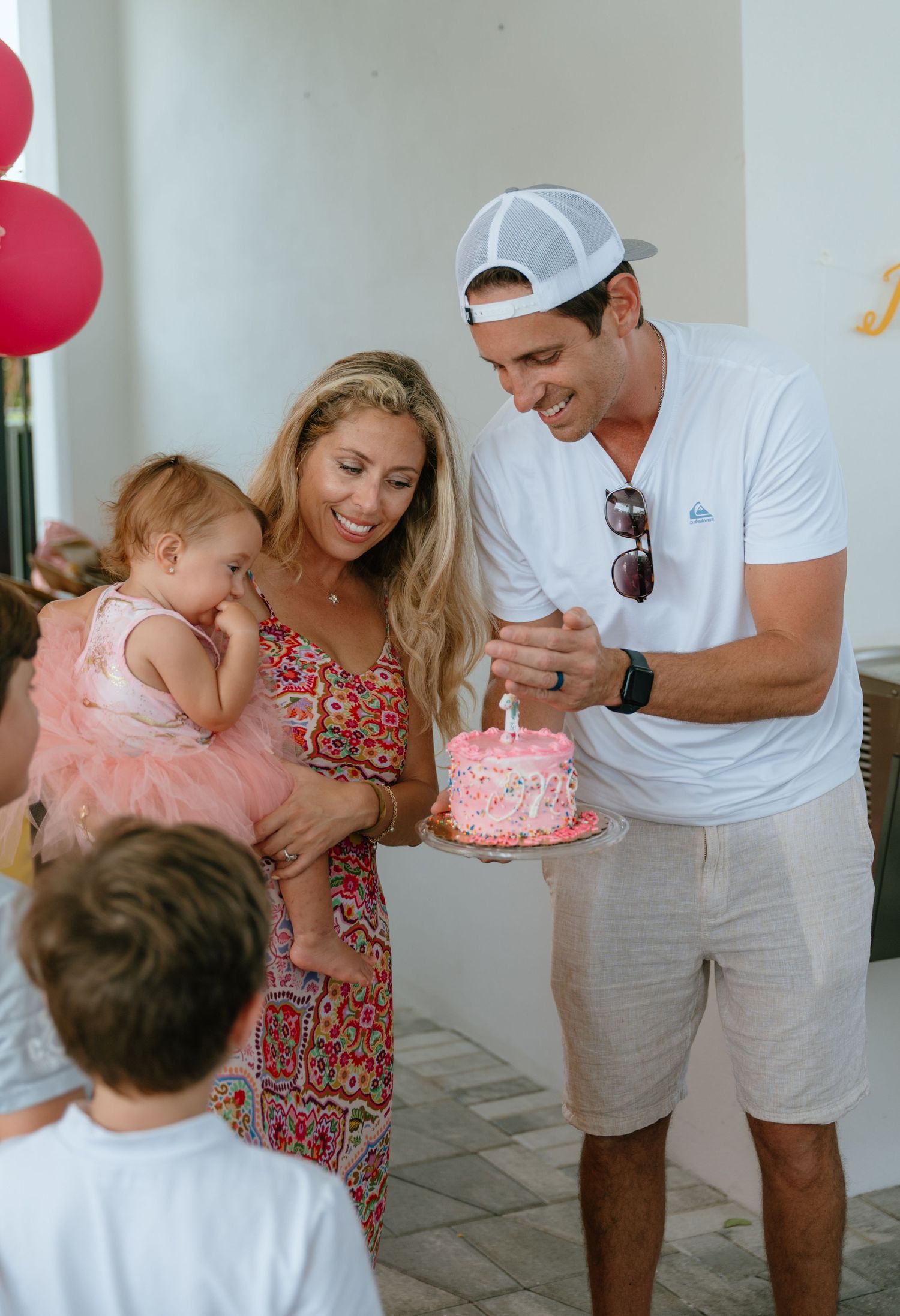 Parents lighting a pink birthday cake candle while holding their baby in a pink tutu at an outdoor birthday party.