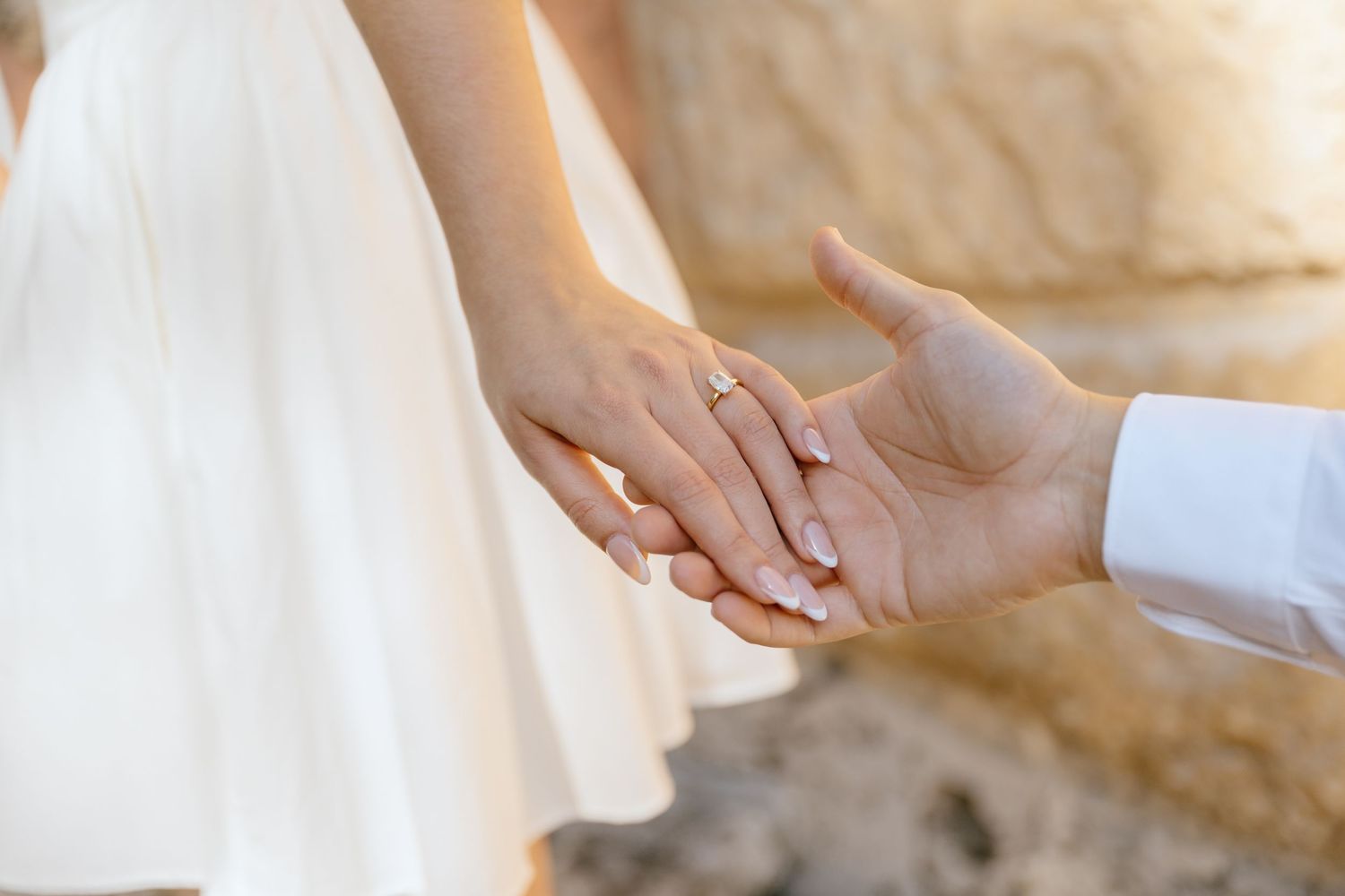 Close-up of couple holding hands showing diamond engagement ring near a stone wall outdoors.