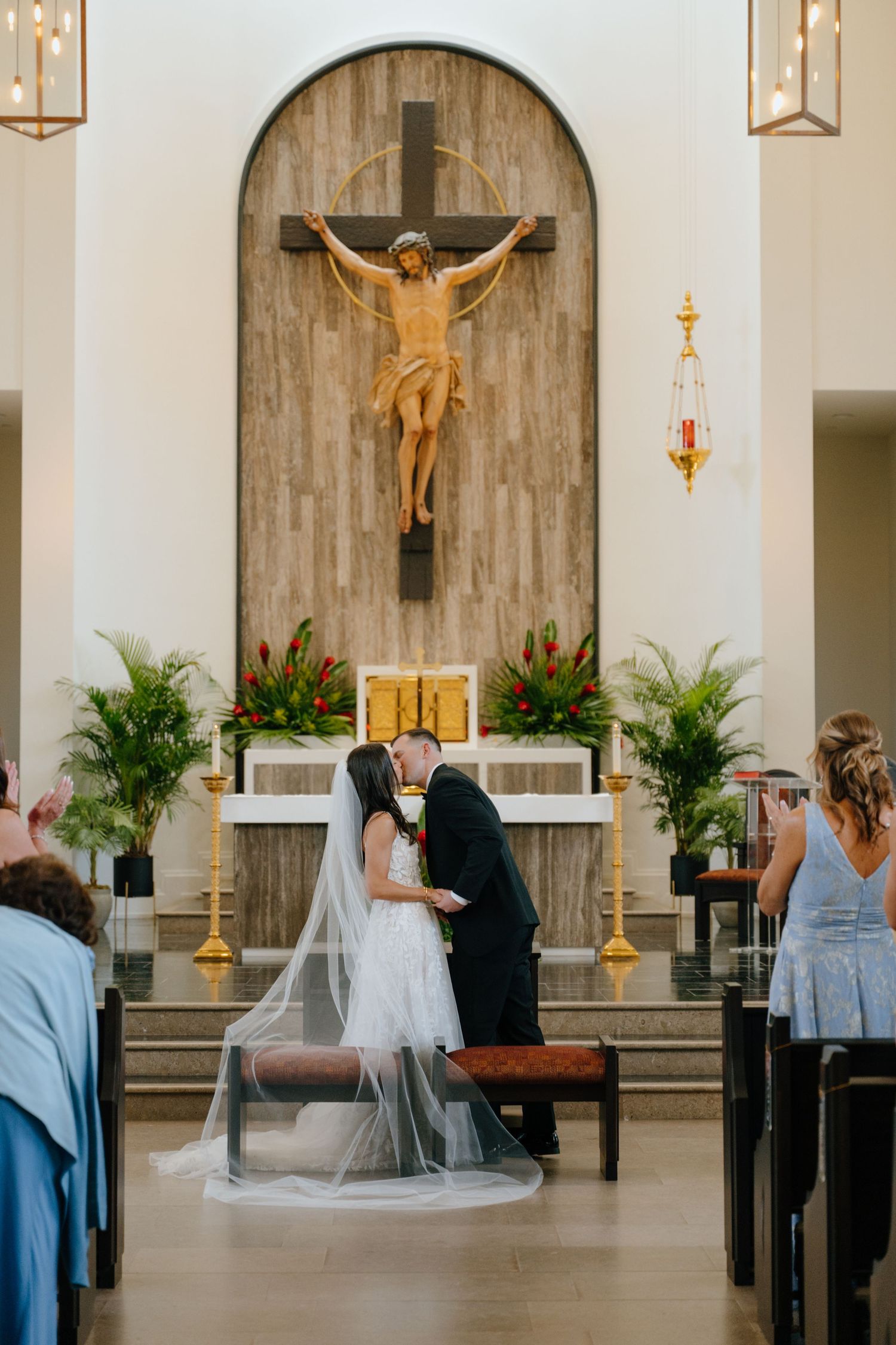 Bride and groom sharing first kiss at the altar beneath a crucifix in a Catholic church.