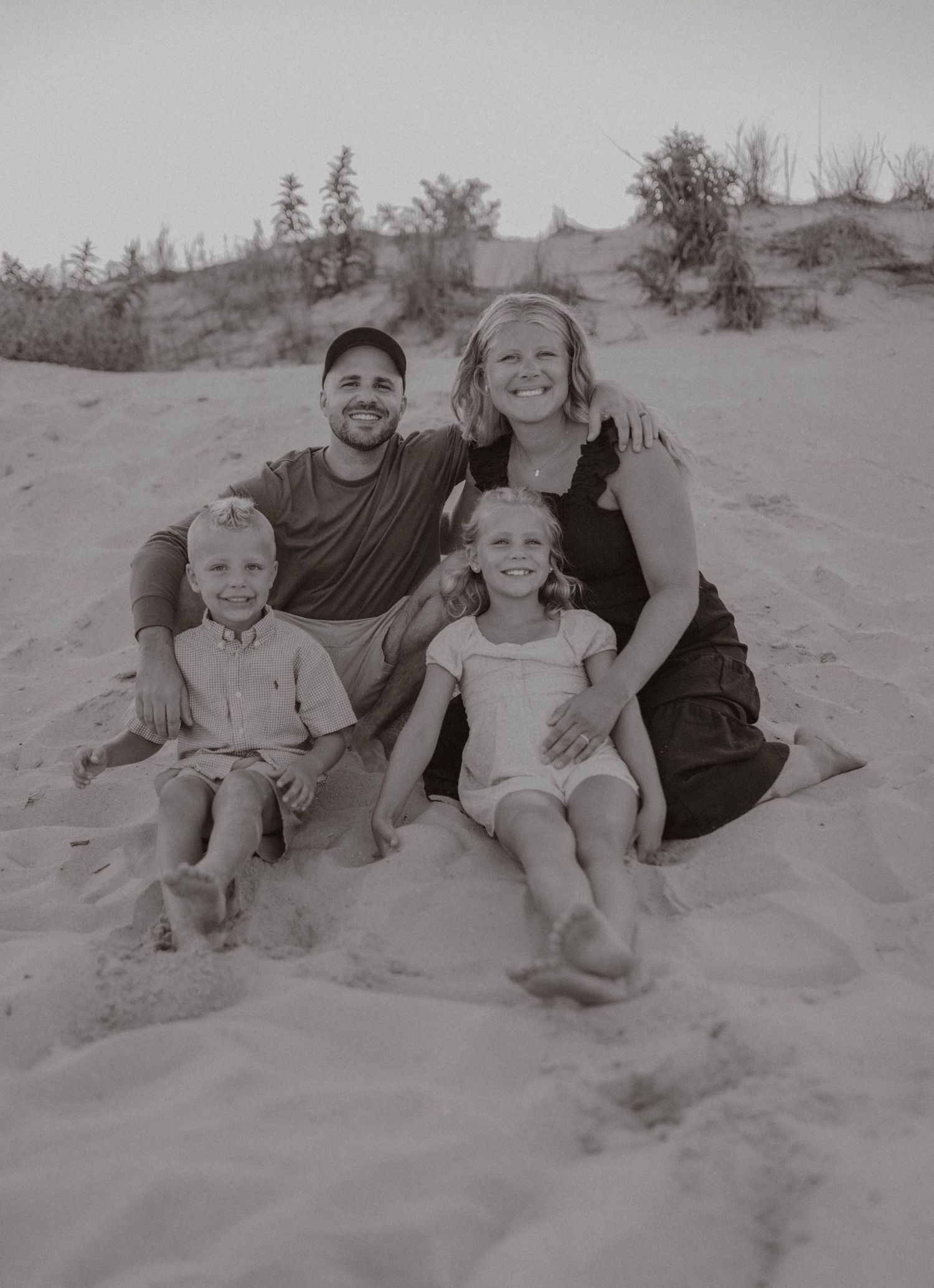 Black and white family portrait of parents and two young children sitting together on sandy dunes.