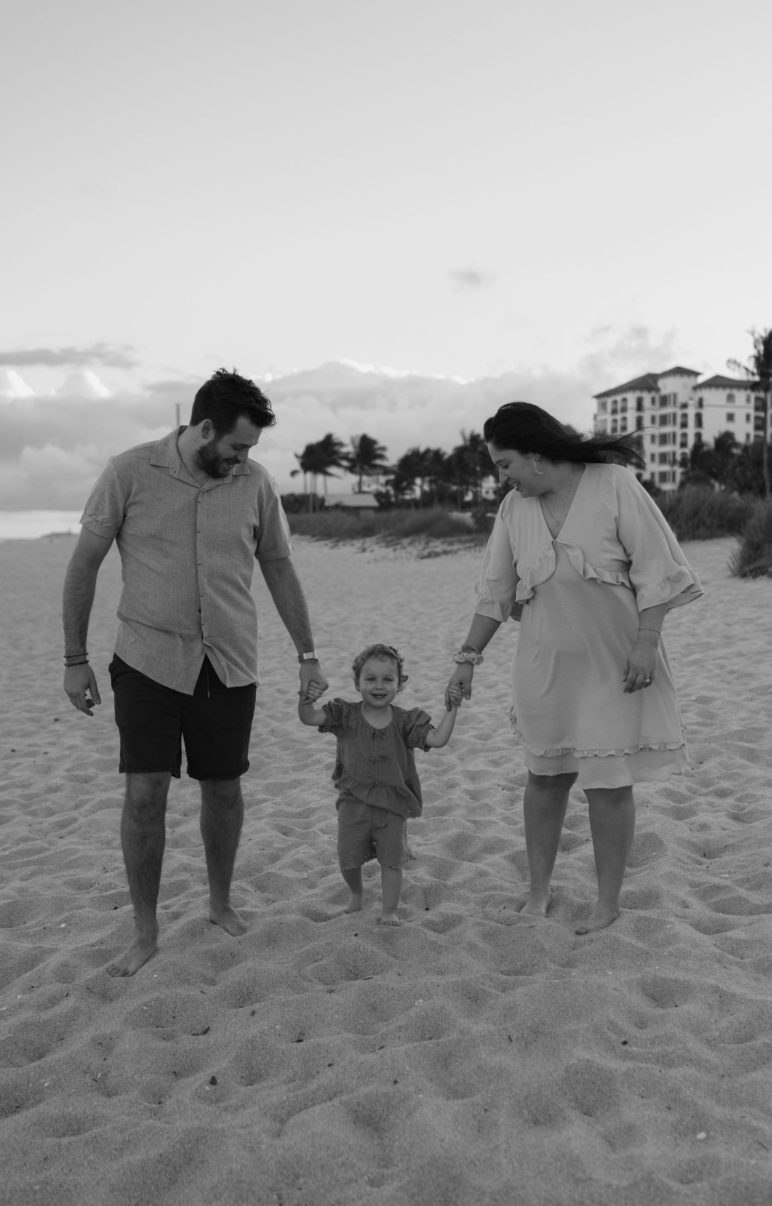 Black and white photo of young parents holding hands with their toddler on a wide sandy beach.