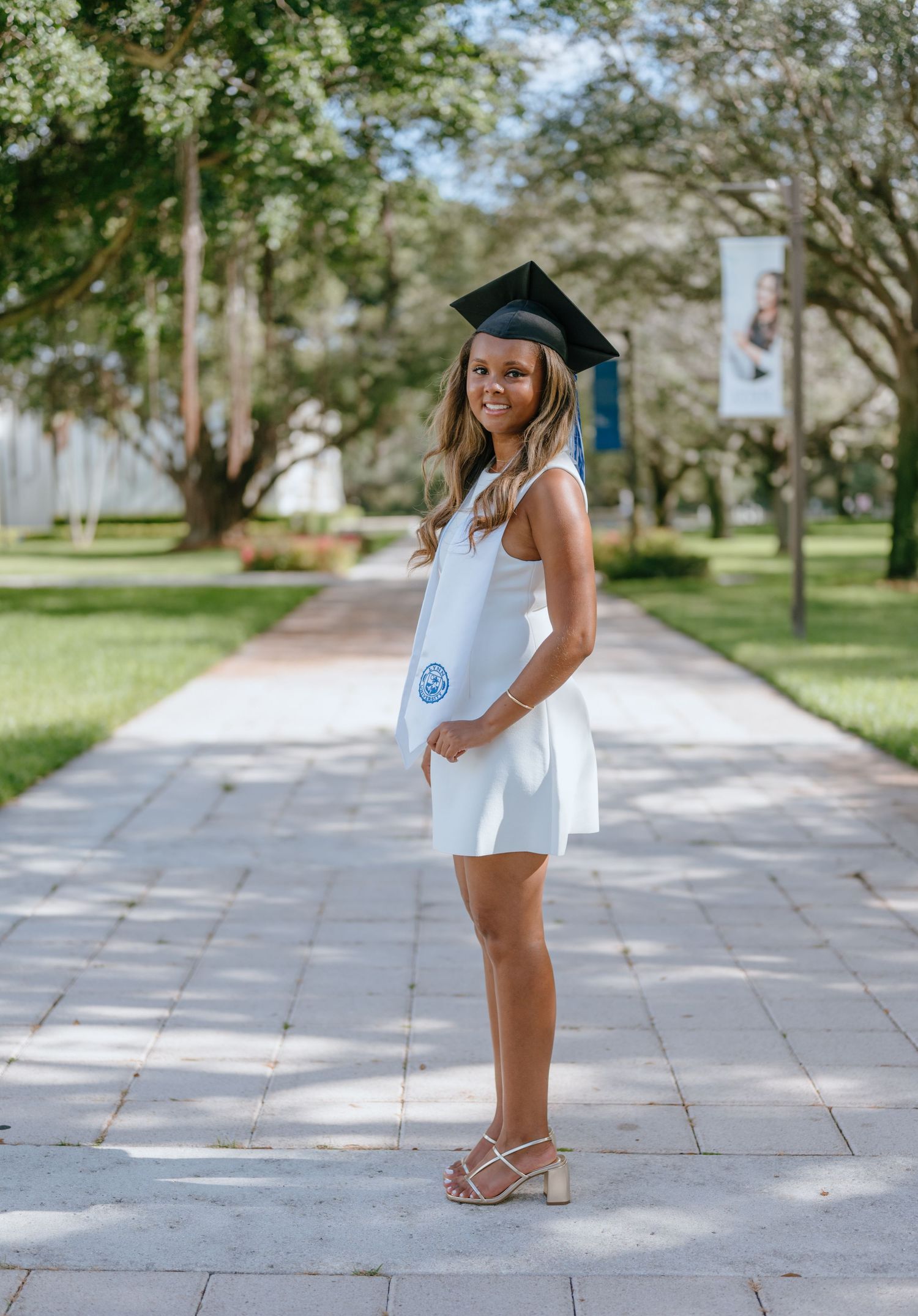 Smiling graduate in white dress and black cap holding diploma on sunny campus walkway.