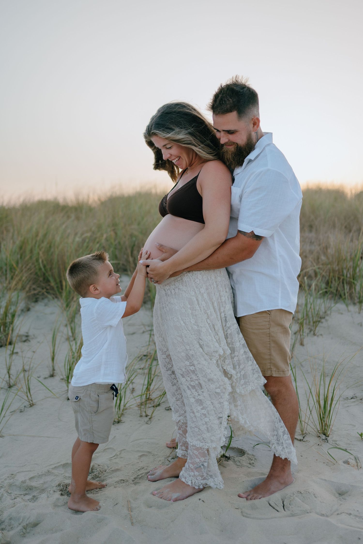 Expecting family with young son touching mom's pregnant belly on sandy beach dunes at sunset.