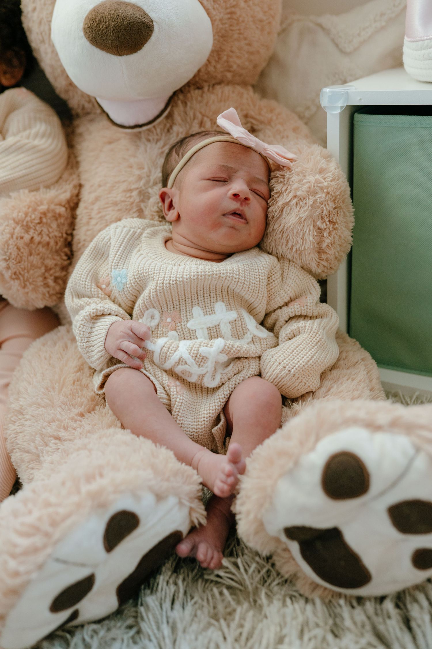 Sleeping newborn baby in a knit romper resting against a large teddy bear.