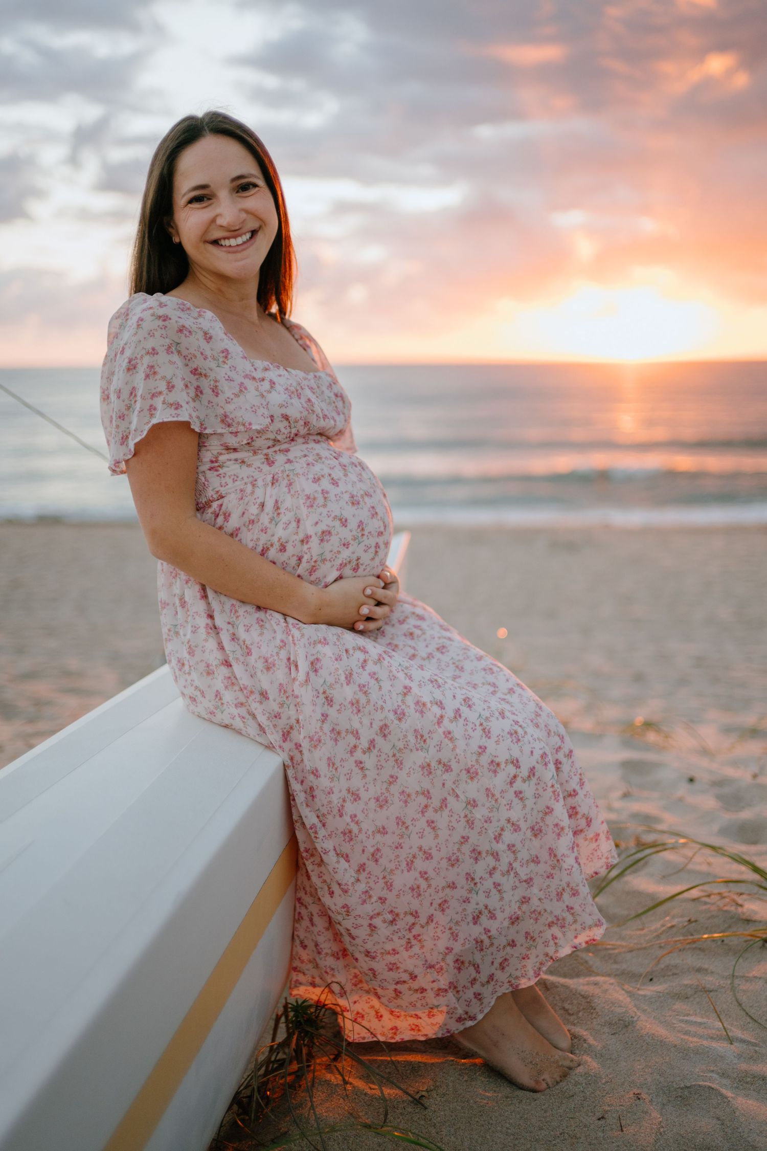 Smiling pregnant woman in floral dress sitting on white bench at colorful beach sunset.