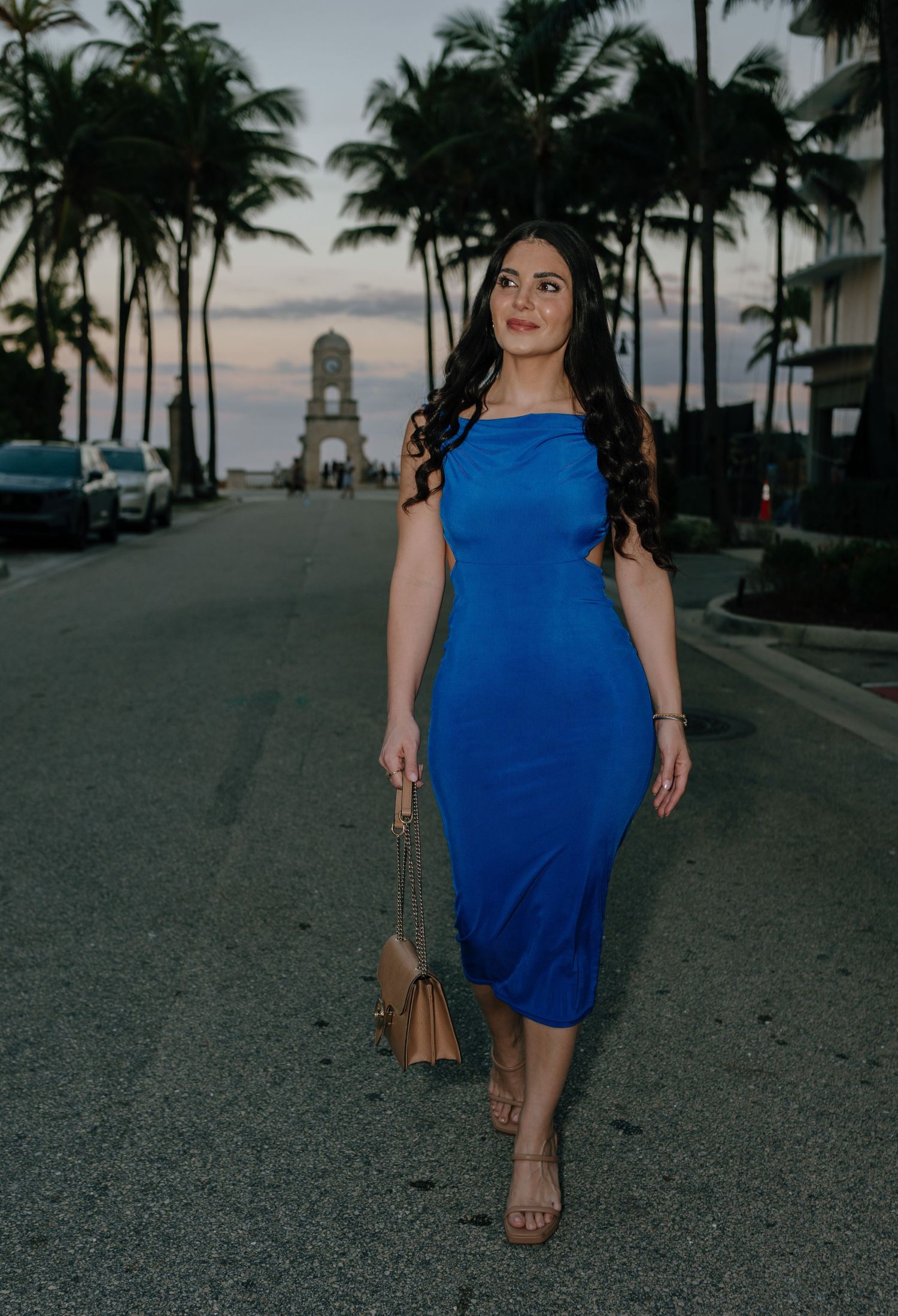 Woman in cobalt blue midi dress strolling palm-lined coastal street at dusk.