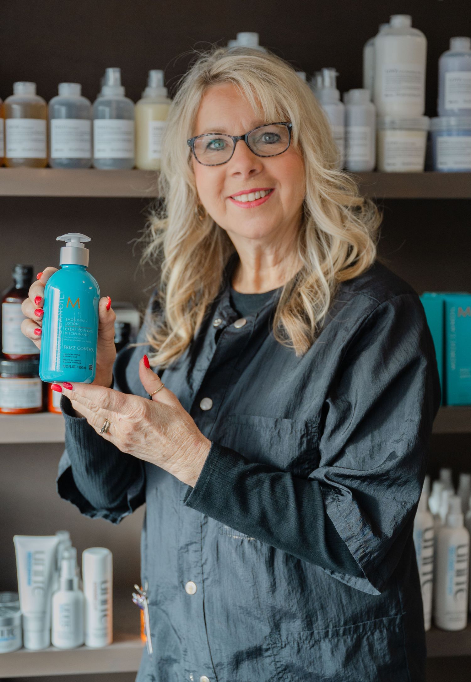 Smiling blonde woman holding Moroccanoil hair product in front of salon shelves.