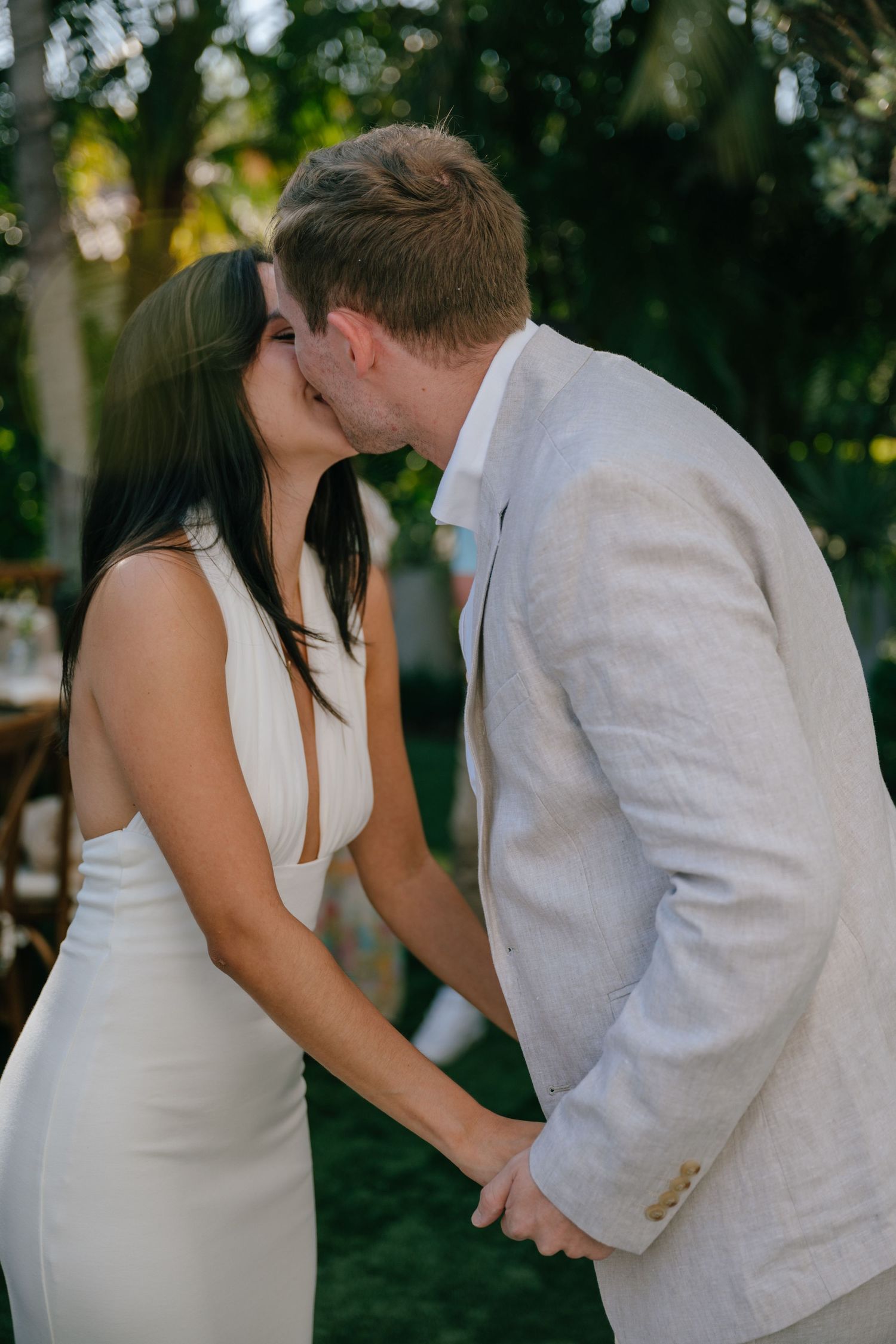 Bride and groom share a romantic first kiss holding hands at an outdoor garden wedding.