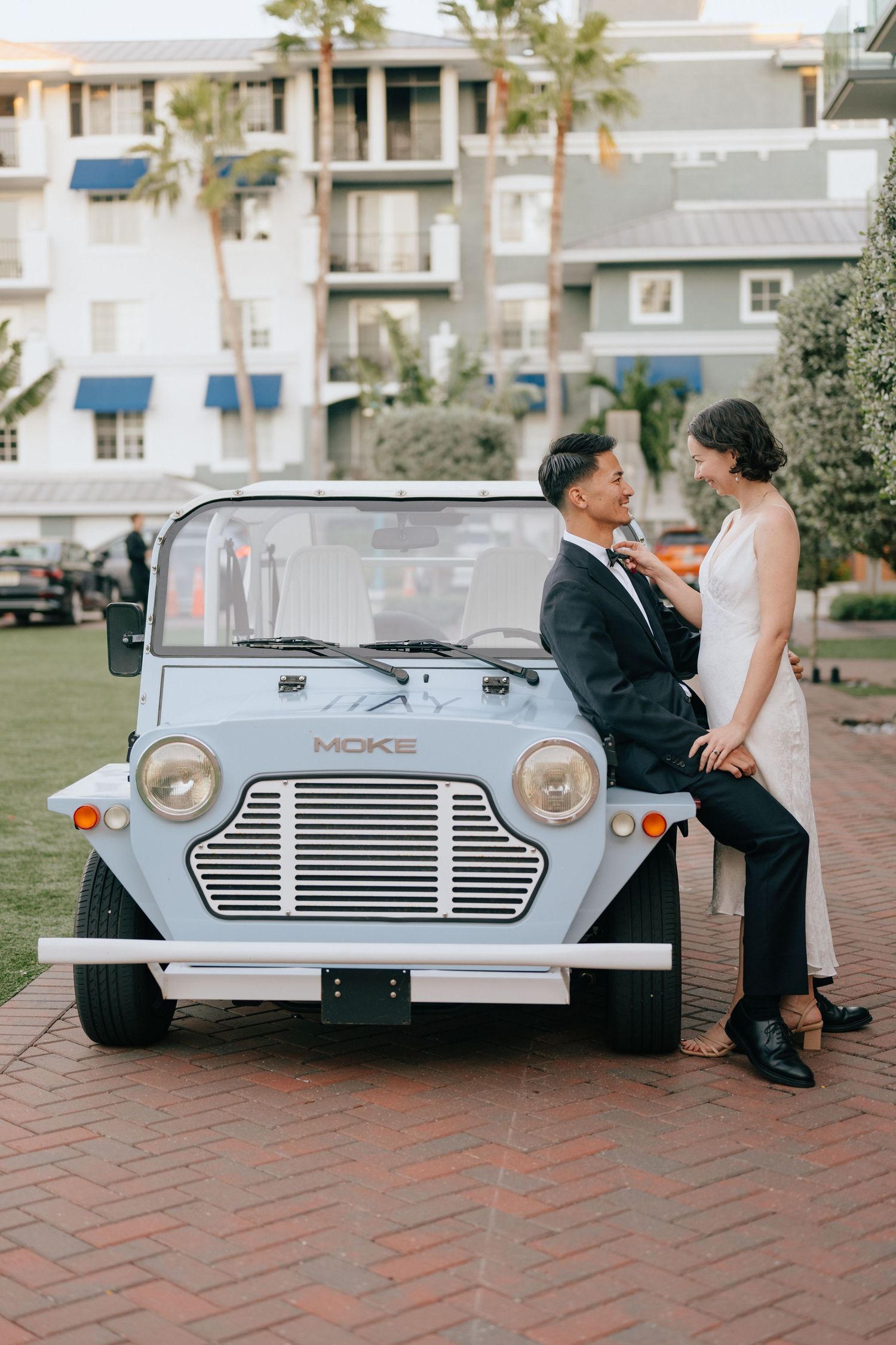 Bride and groom posing with a vintage light blue Moke car on brick pavement outside a resort.