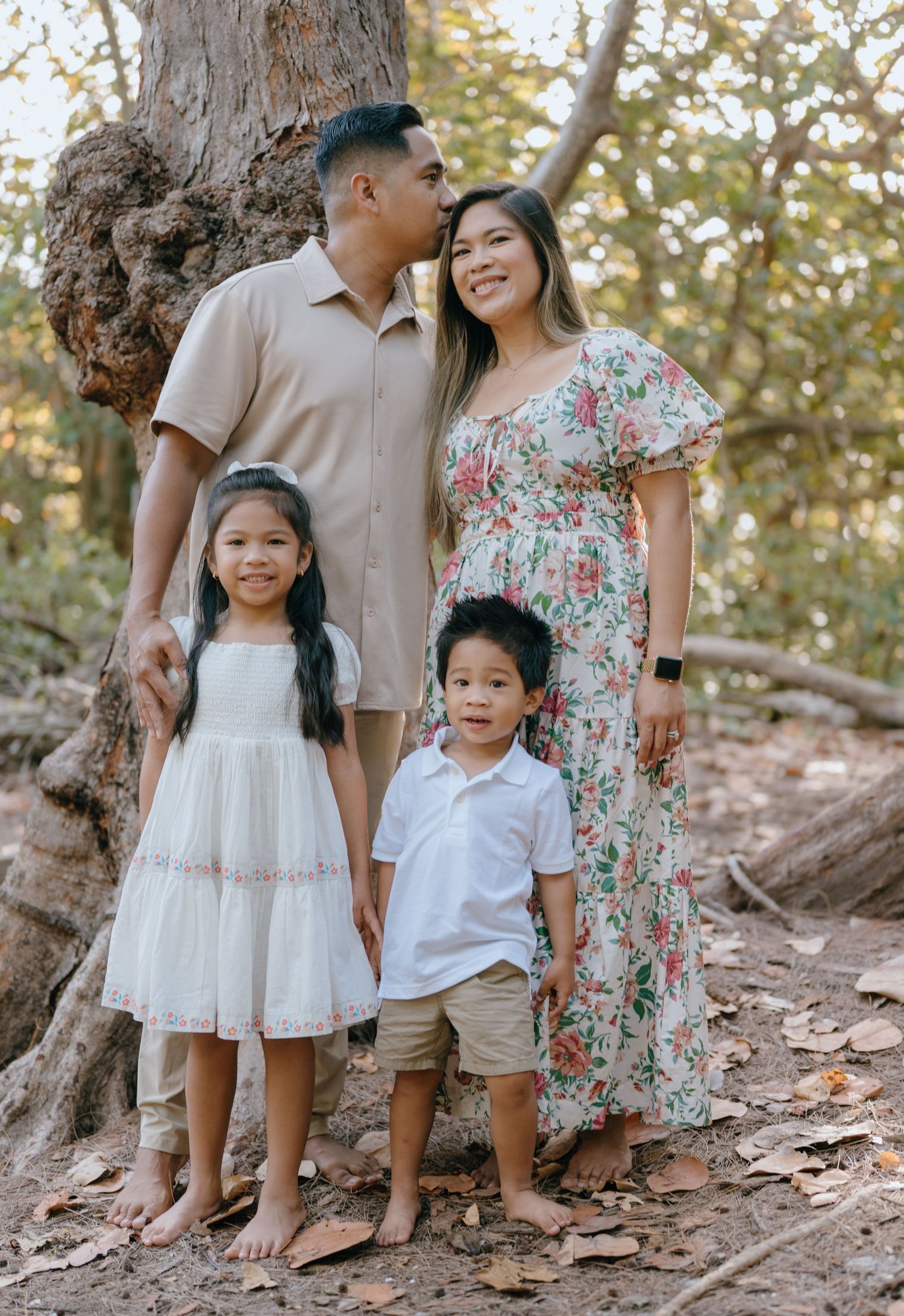Asian family of four posing warmly near a tree in a sunlit outdoor setting.