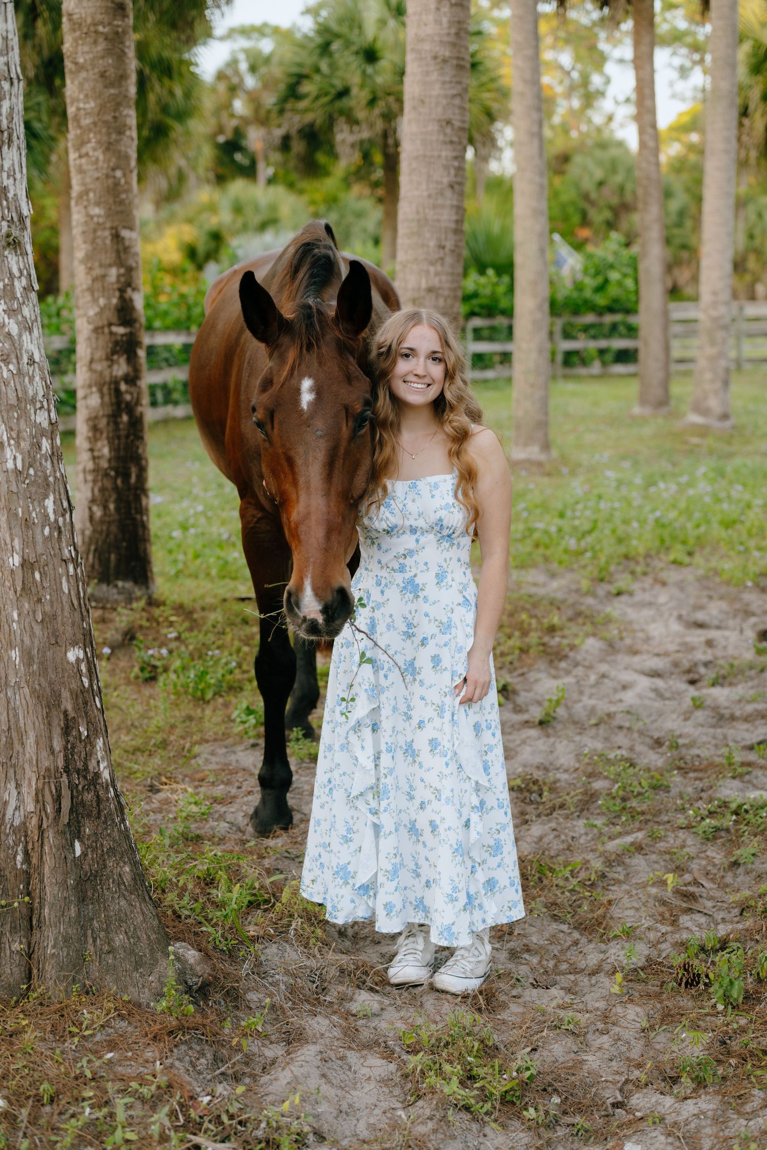 A smiling teenage girl in a white floral dress stands beside a bay horse among palm trees.