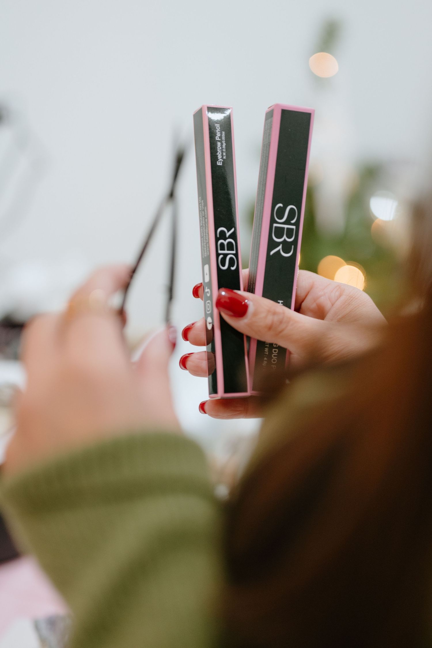 Close-up of hands with red nails holding two SBR branded eyebrow product boxes near a festive holiday display.