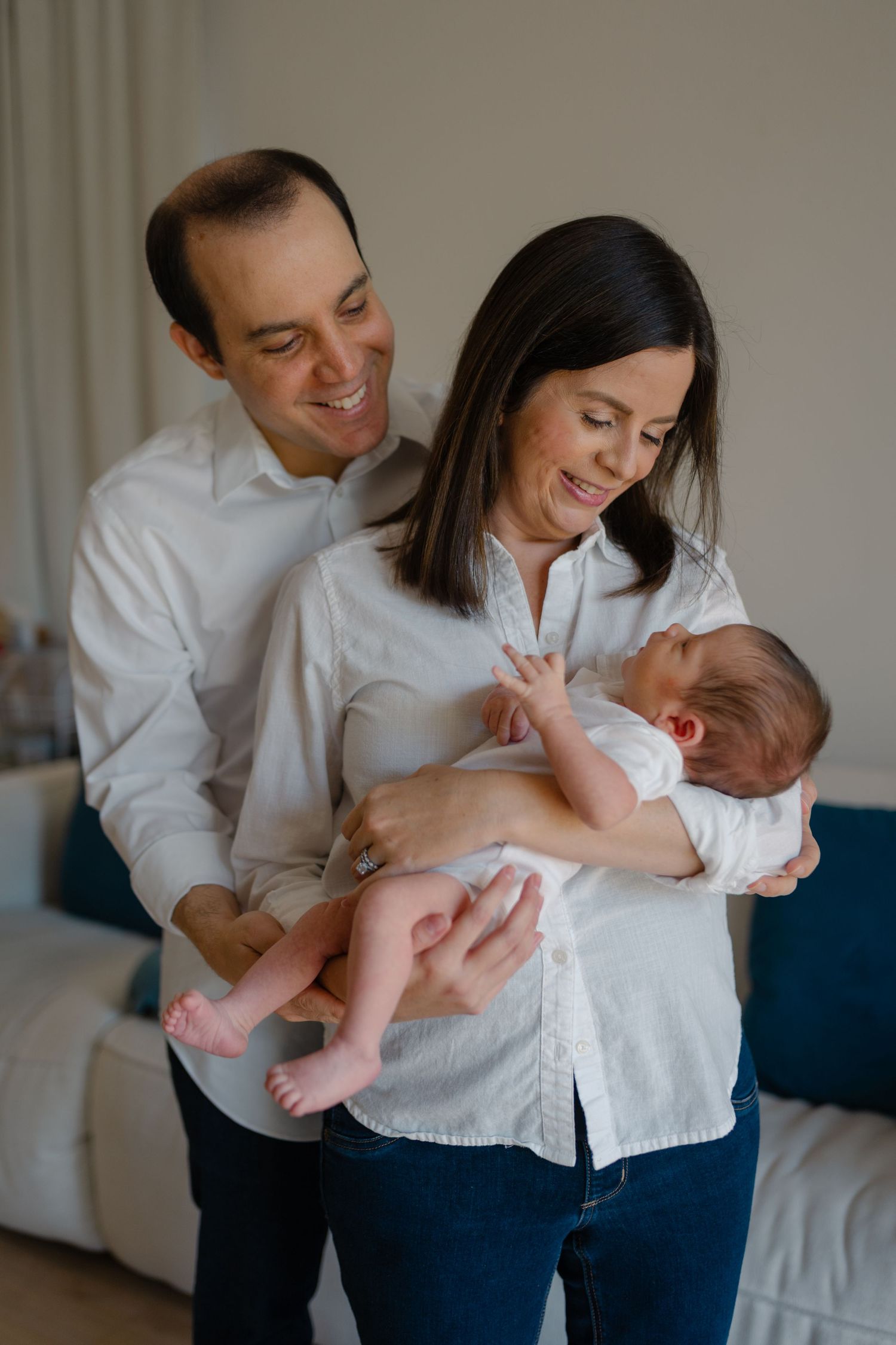 Smiling parents in white shirts lovingly hold their newborn baby at home.