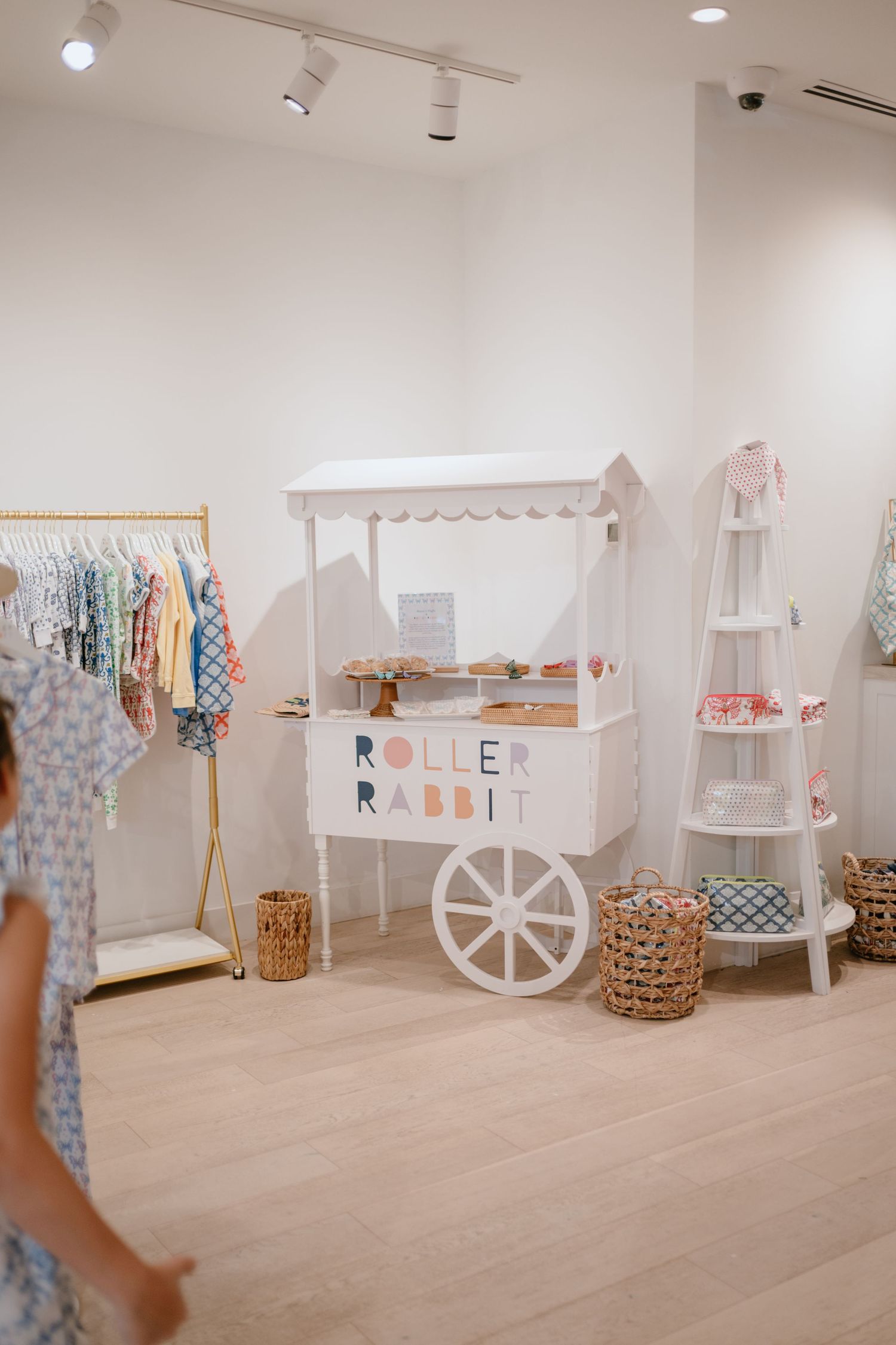 Roller Rabbit boutique store interior with white cart display, clothing rack, and wicker baskets on light wood floors.