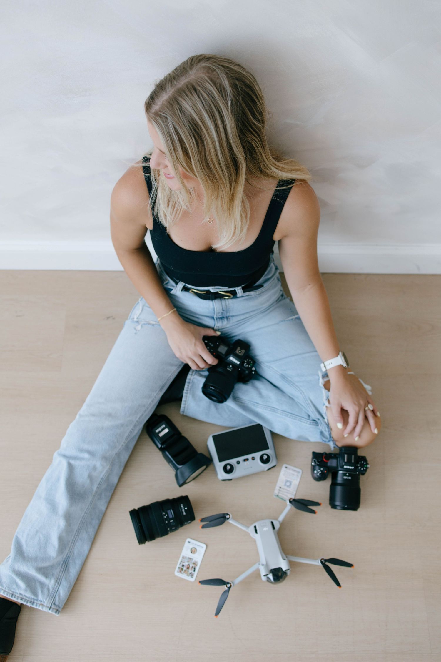 Blonde female photographer sitting on the floor surrounded by cameras, drone, and photography equipment.