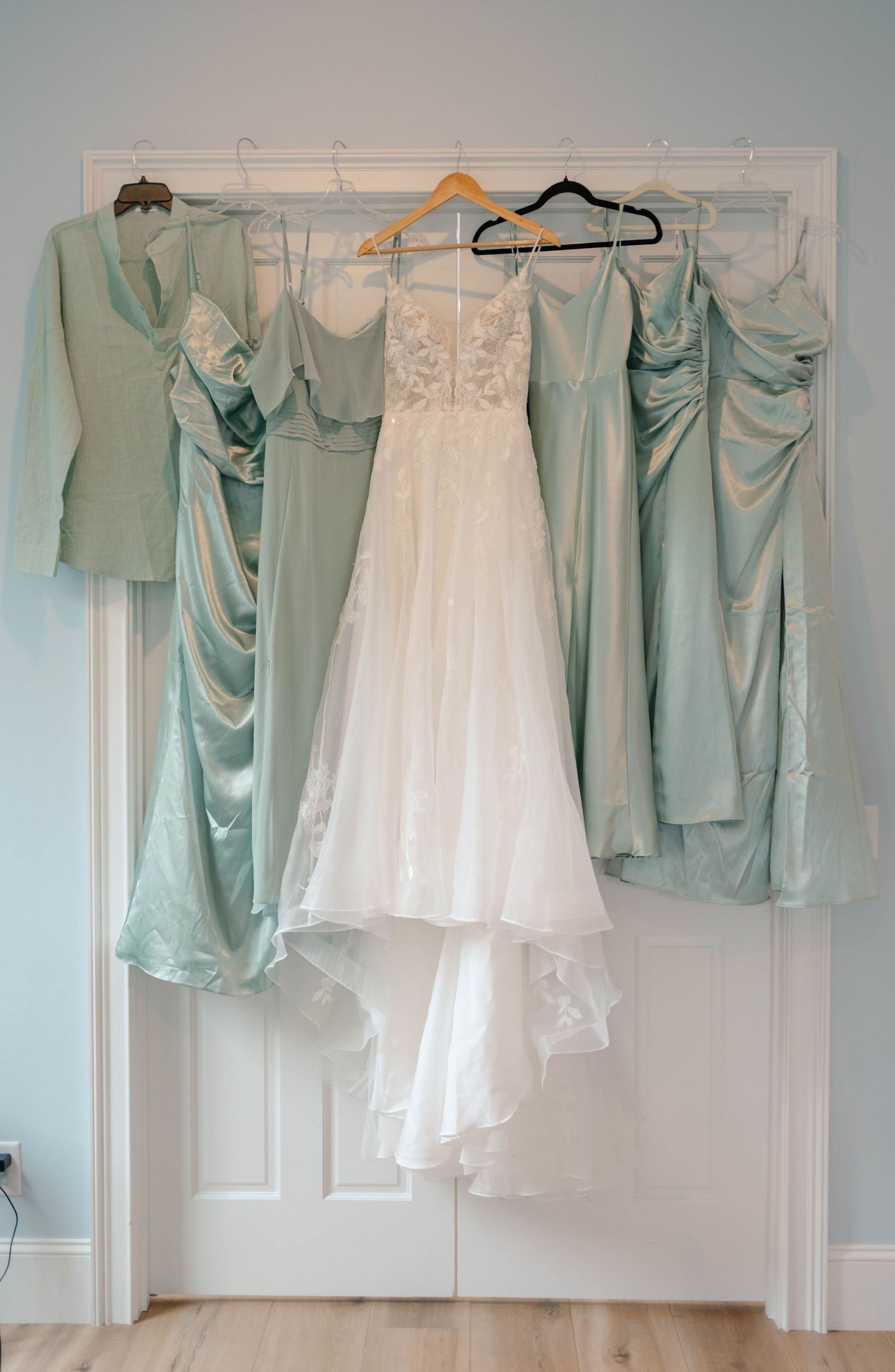 White lace wedding gown hanging between sage green satin bridesmaid dresses on a white door.