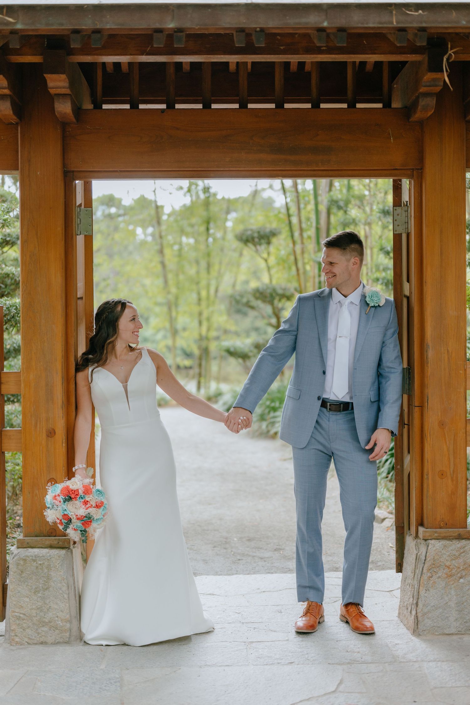 Smiling bride and groom hold hands under a wooden gazebo arch with greenery behind them.