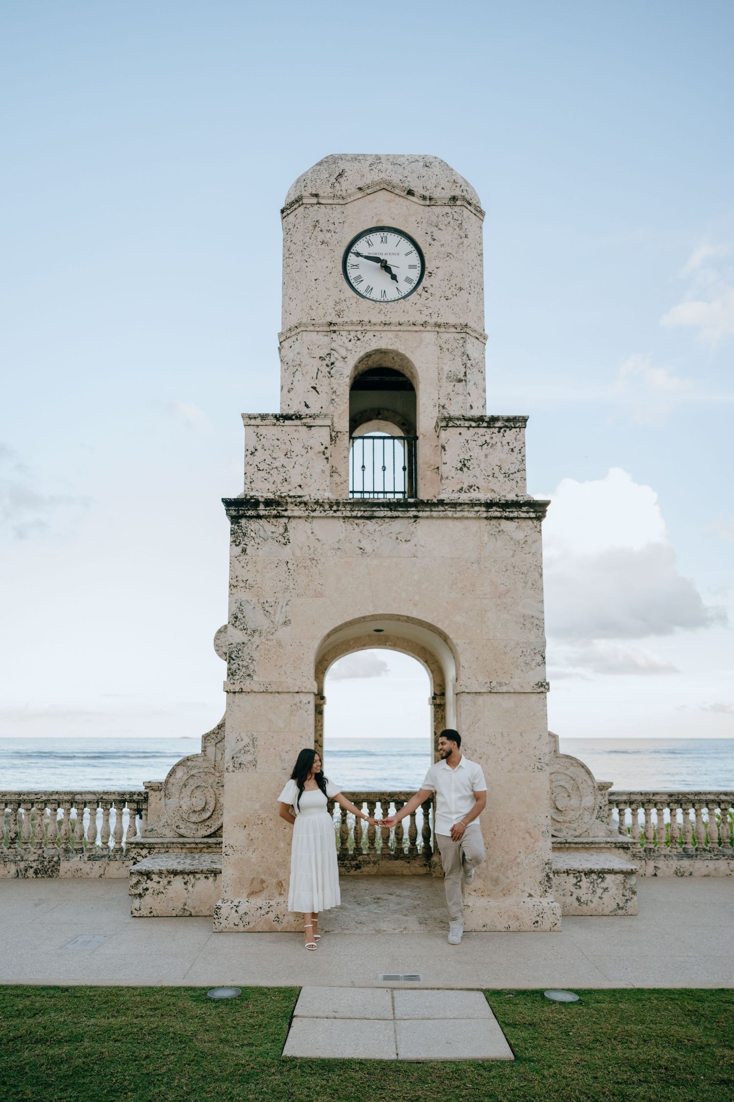 Couple in white outfits standing beneath a historic stone clock tower by the ocean.