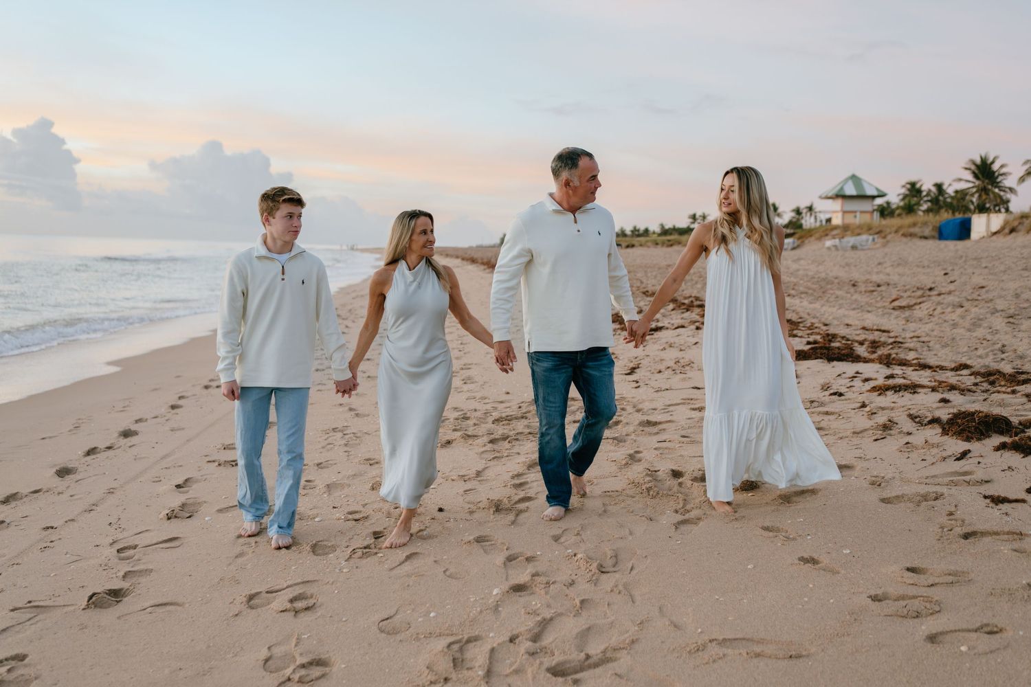 A family of four walks hand-in-hand along a sandy beach at dusk.