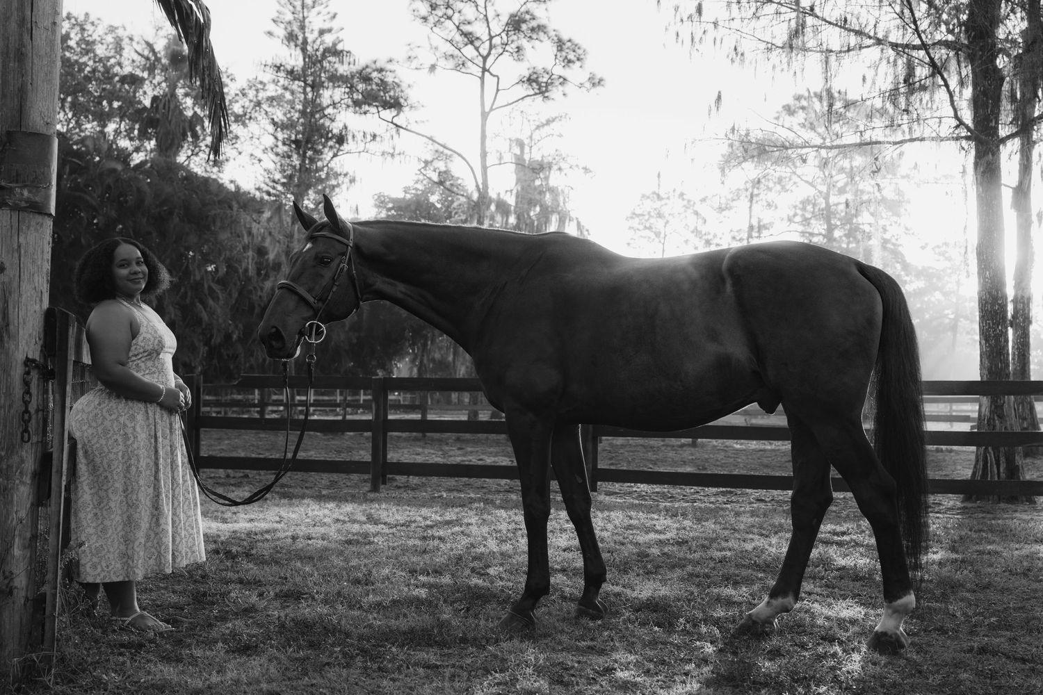 Black and white photo of a woman standing beside a tall dark horse in a sunlit paddock.