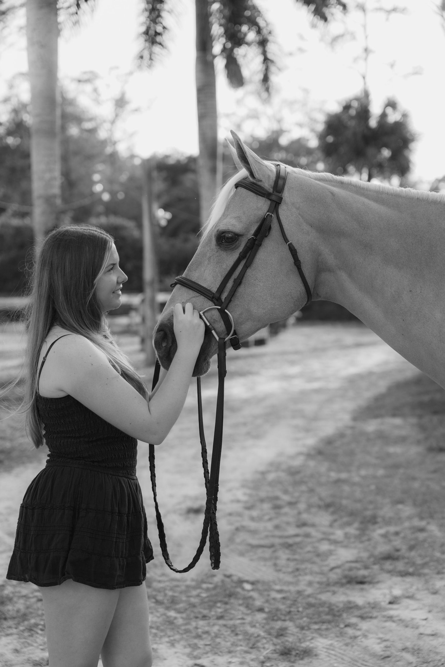 Young woman in black dress gently holds face of palomino horse outdoors, black and white equestrian photo.