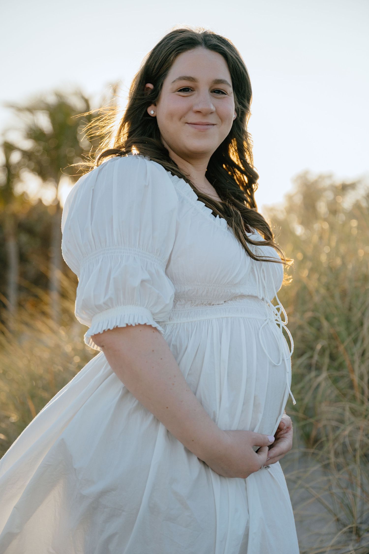 Pregnant woman in white dress cradles baby bump while standing in golden sunset dunes.