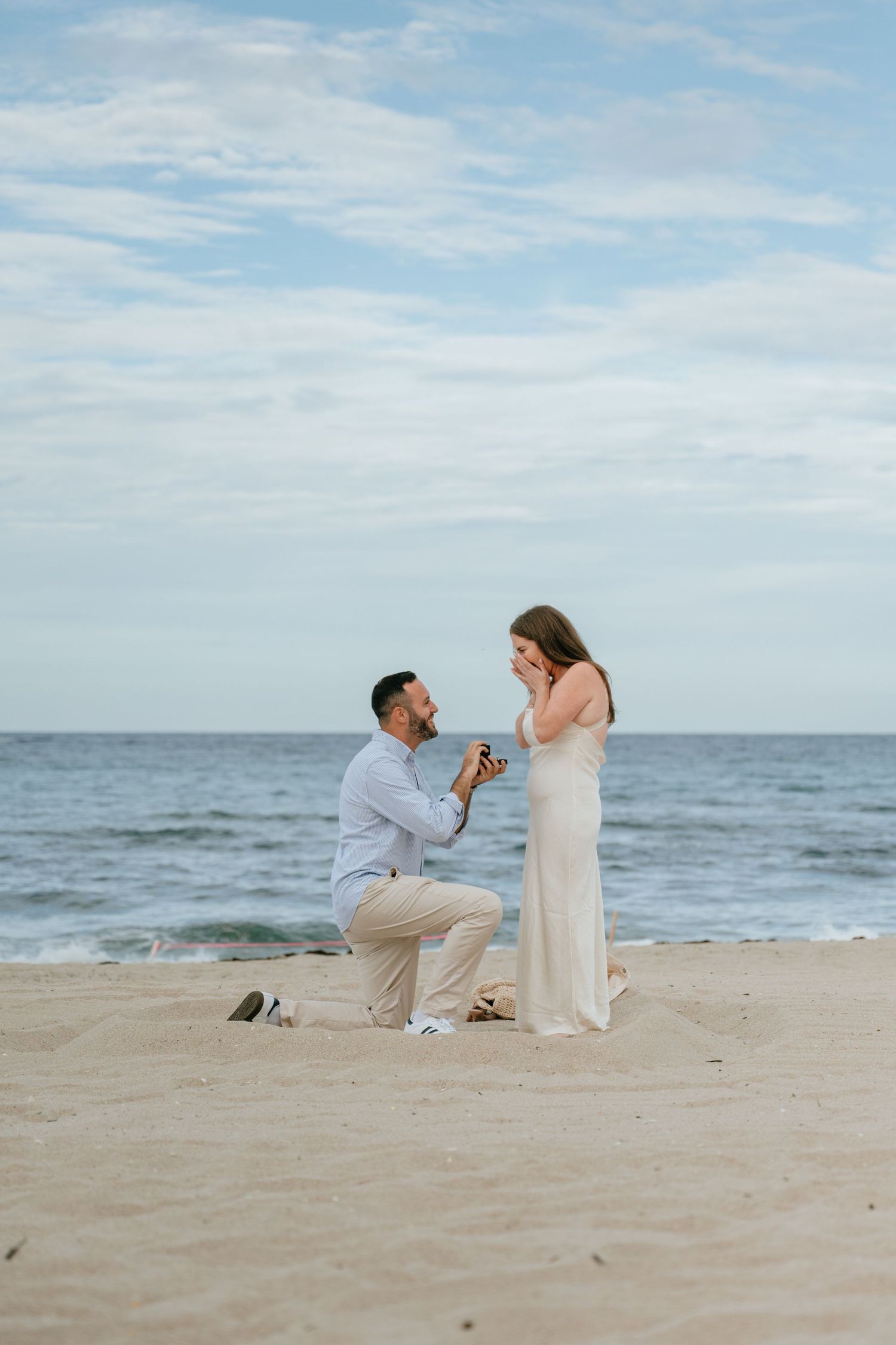 Romantic beach marriage proposal with man on one knee presenting ring to emotional woman in white dress.